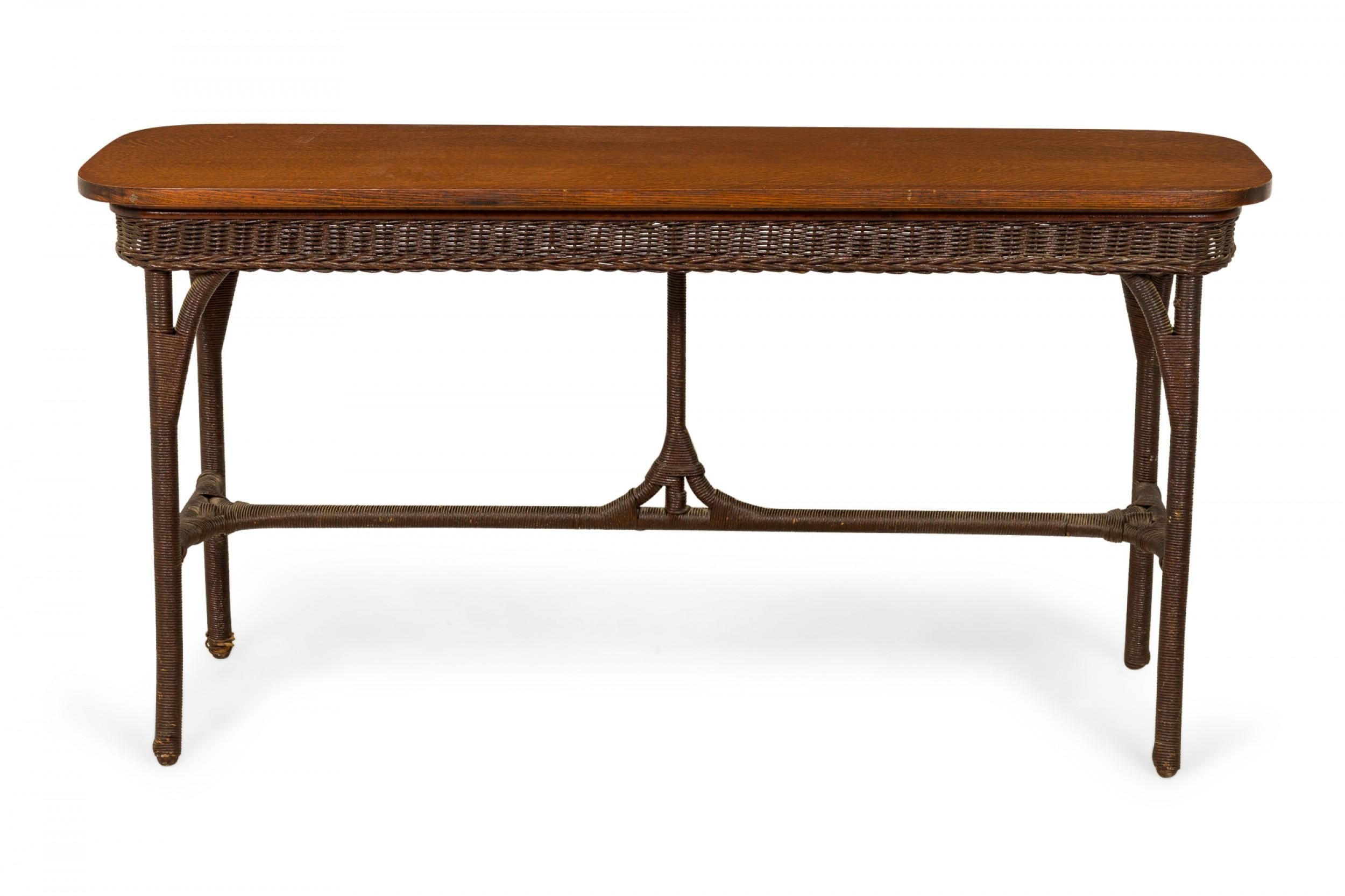 American wicker (1940s) and oak console / davenport table with a rectangular solid oak top with rounded corners atop a dark brown painted wicker-wrapped base with a decorative wicker apron, sabre legs, and a stretcher base with central support.