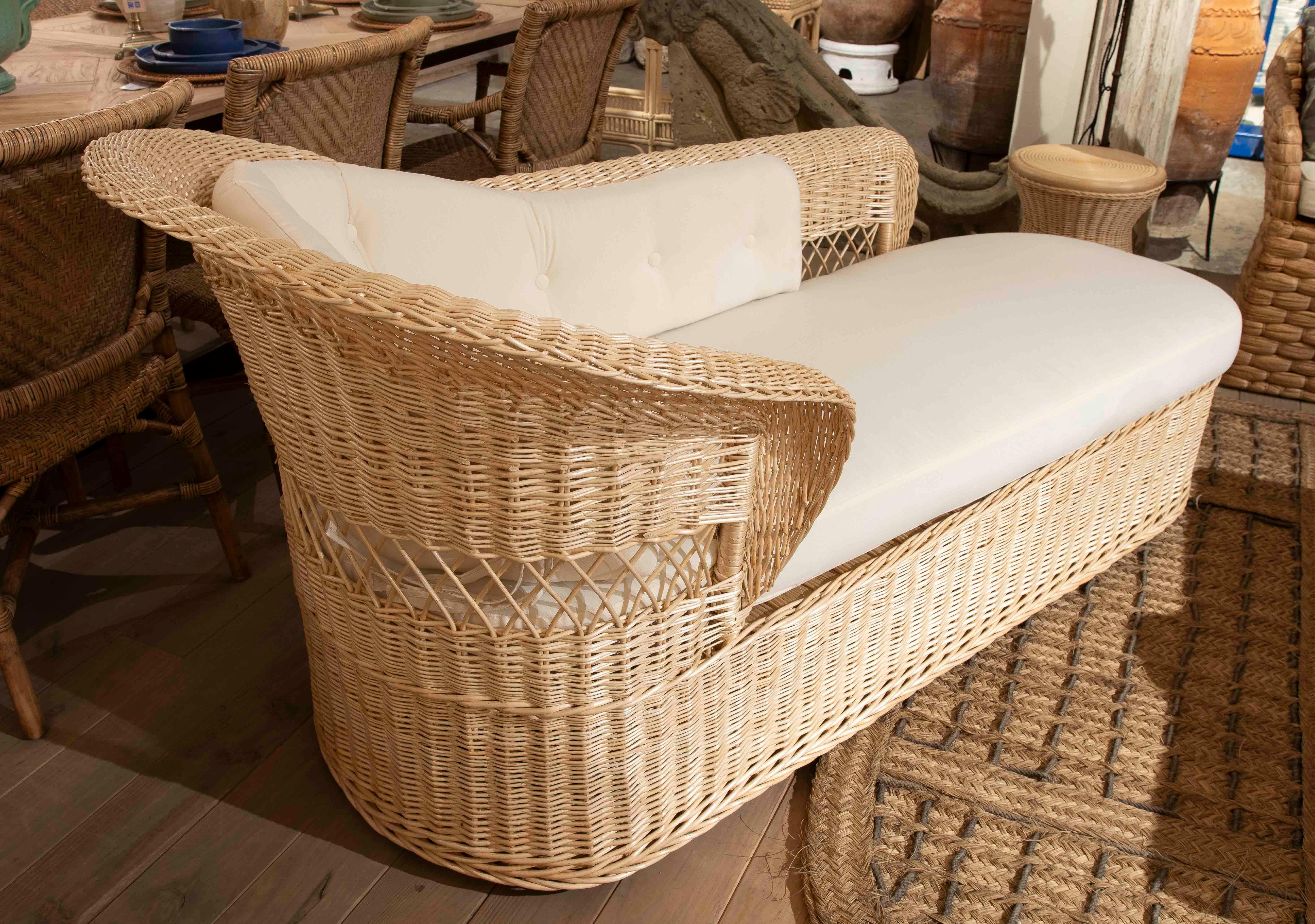 Elegante chaise longue in vimini intrecciato a mano, con un design avvolgente che unisce comfort e raffinatezza. La sua struttura curva e lo schienale imbottito con cuscini bianchi offrono un tocco rilassante e naturale, ideale per gli spazi di