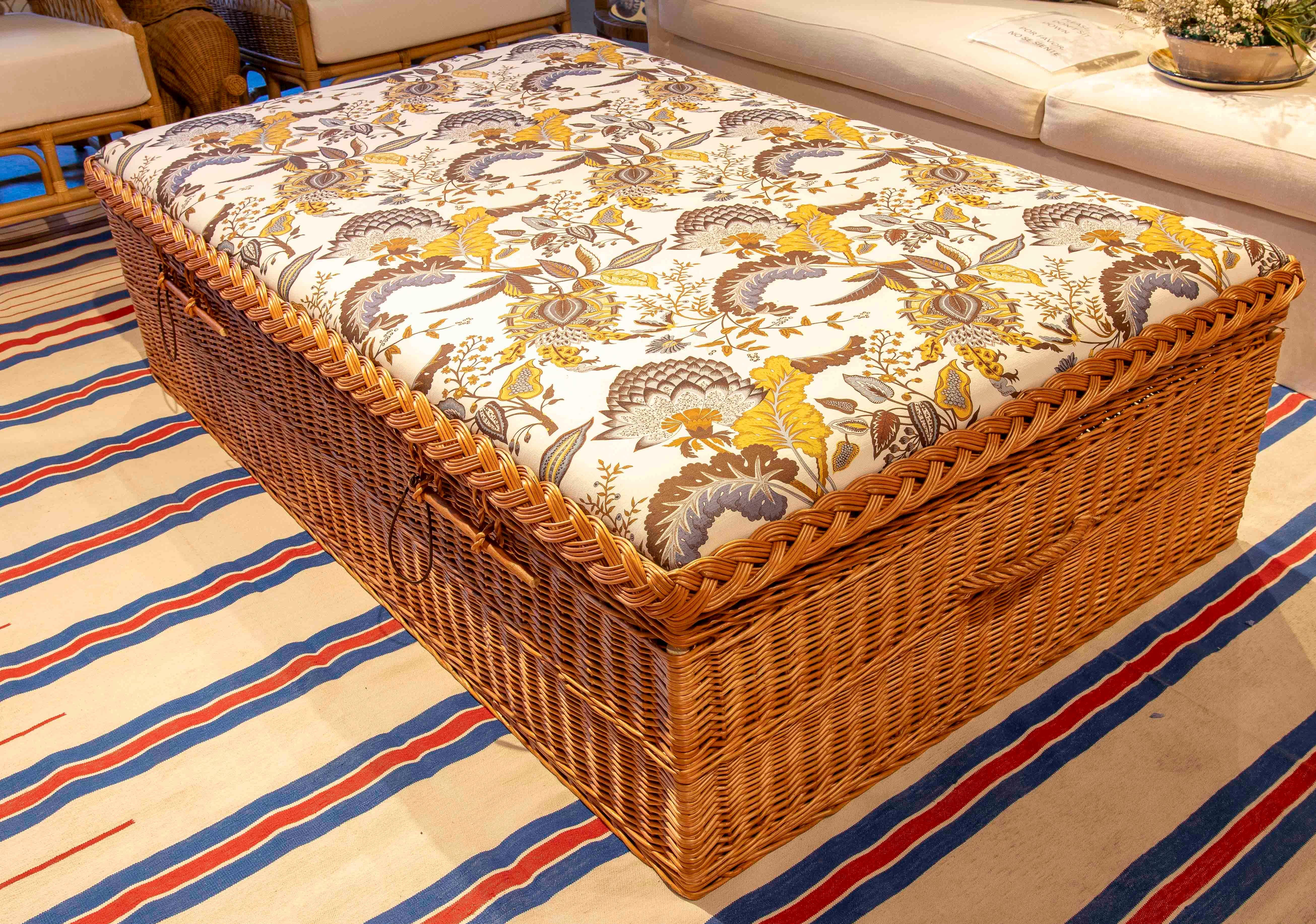 Encantador baúl otomano elaborado en resistente mimbre tejido a mano, con tapa acolchada que funciona como asiento o mesa auxiliar. El cojín superior presenta una tela con estampado floral en tonos mostaza, gris y marrón sobre fondo blanco,