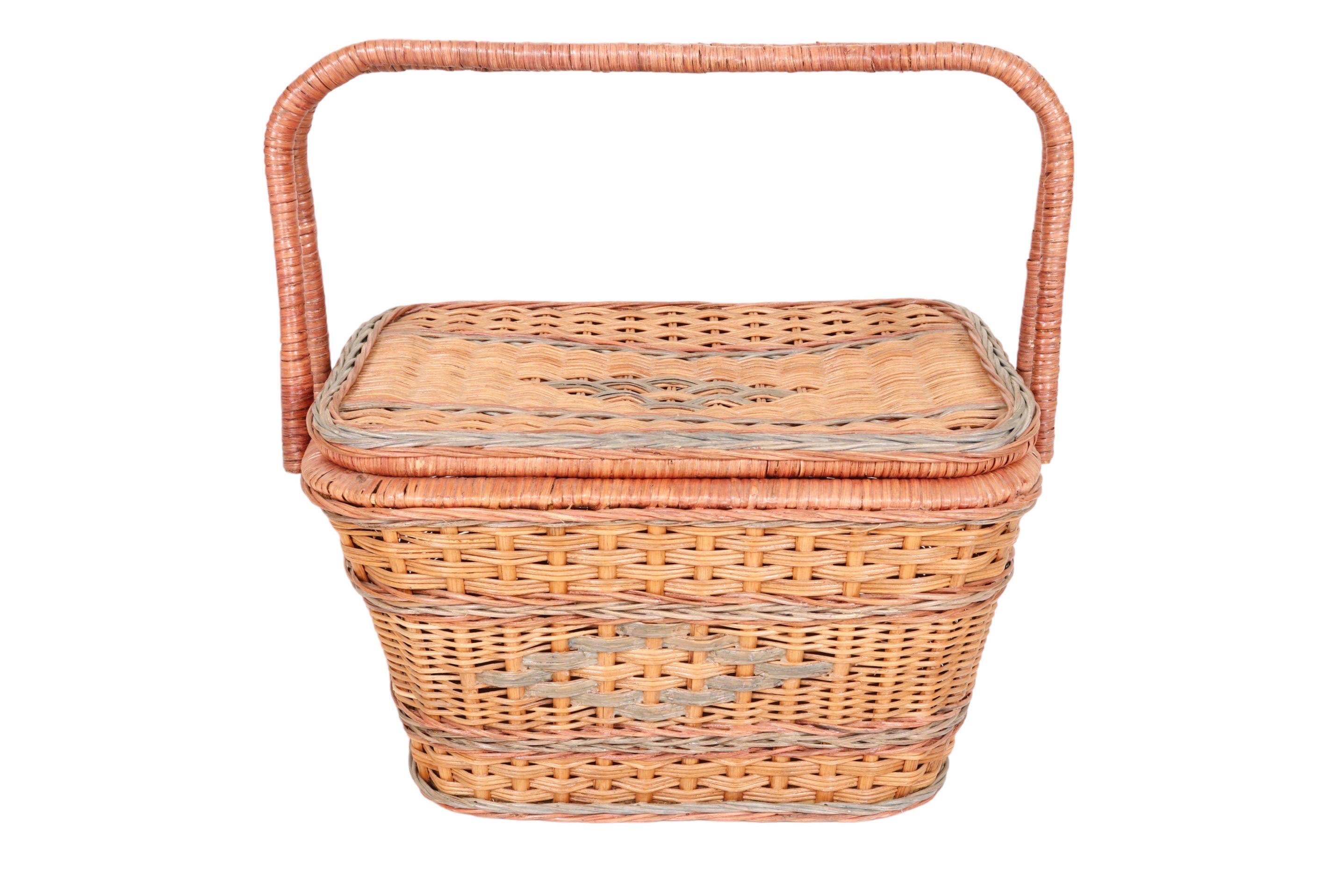 A wicker picnic hamper basket with two wrapped bamboo handles. Decorated on each side with twisted bands of rattan in pink and green, and green woven diamonds reminiscent of Heywood Wakefield. The lid is hinged at one side and opens to reveal plenty