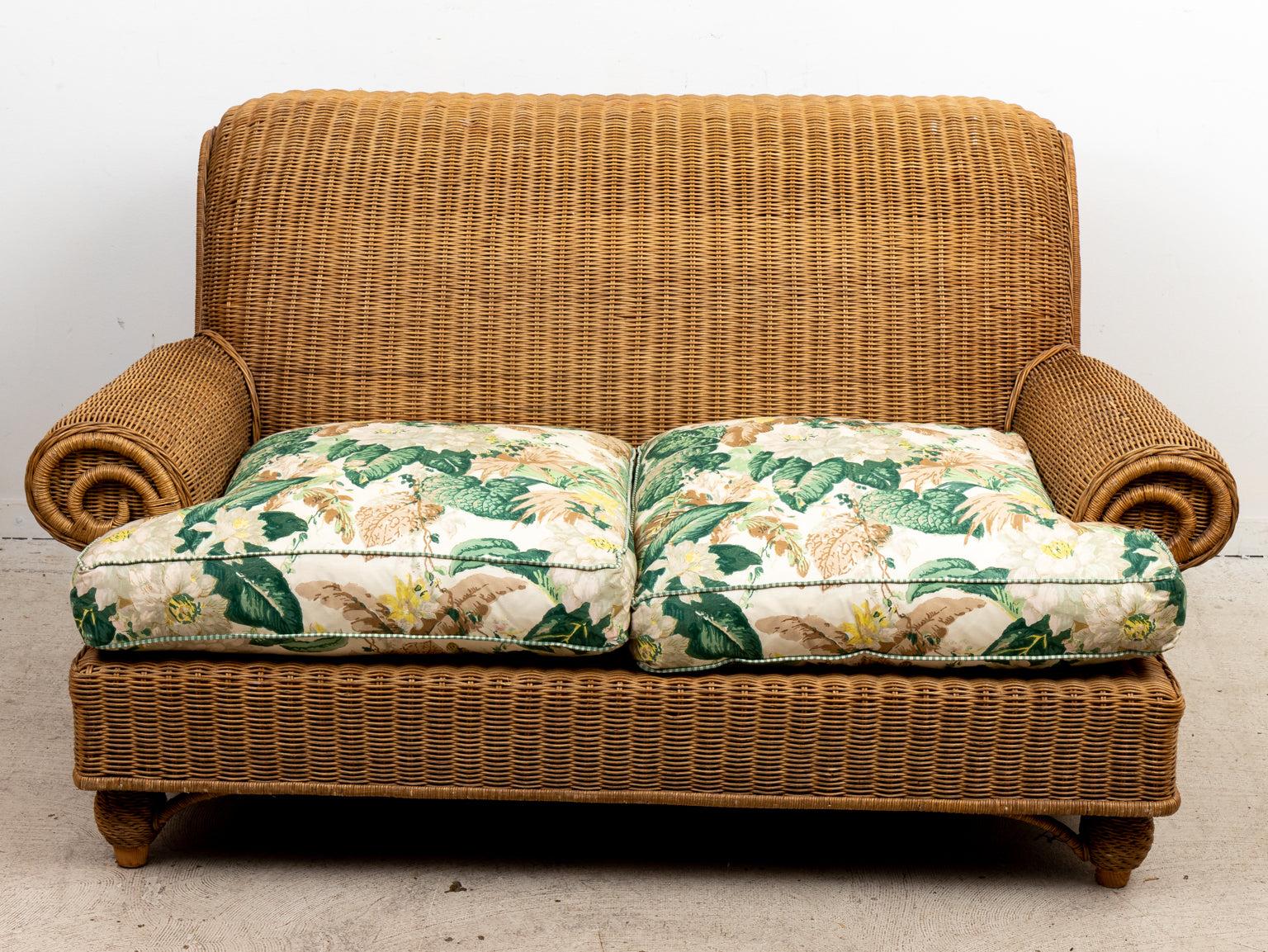 Wicker two-seat sofa with hard rolled arms and back. Two floral upholstered seat cushions and two throw pillows included. Please note of wear consistent with age.