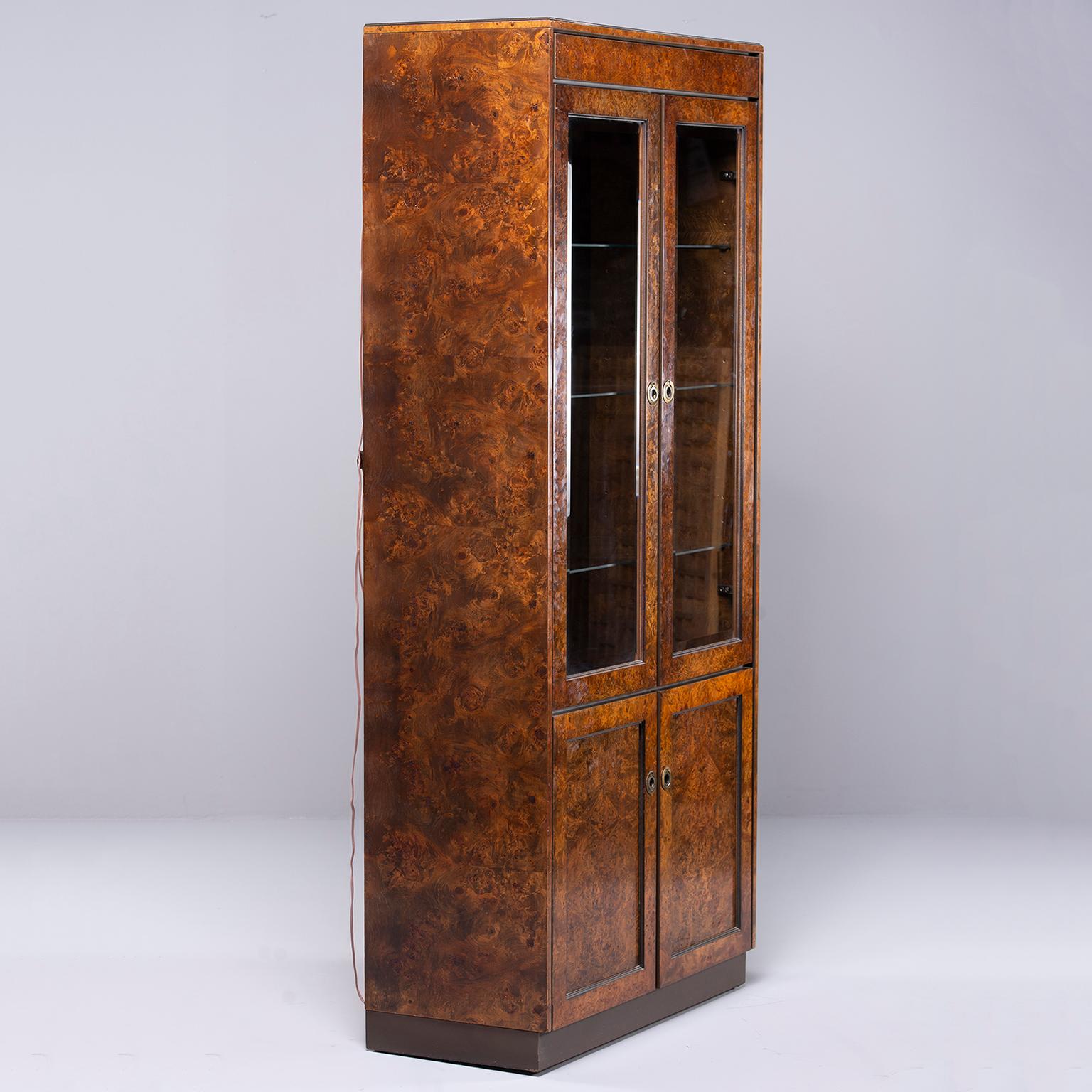 Widdicomb dark burled olive wood cabinet has lower storage compartment with two hinged wood doors, circa 1970s. Top compartment features beveled edge glass front hinged doors and glass shelves with interior light on inside of top. Dark brass