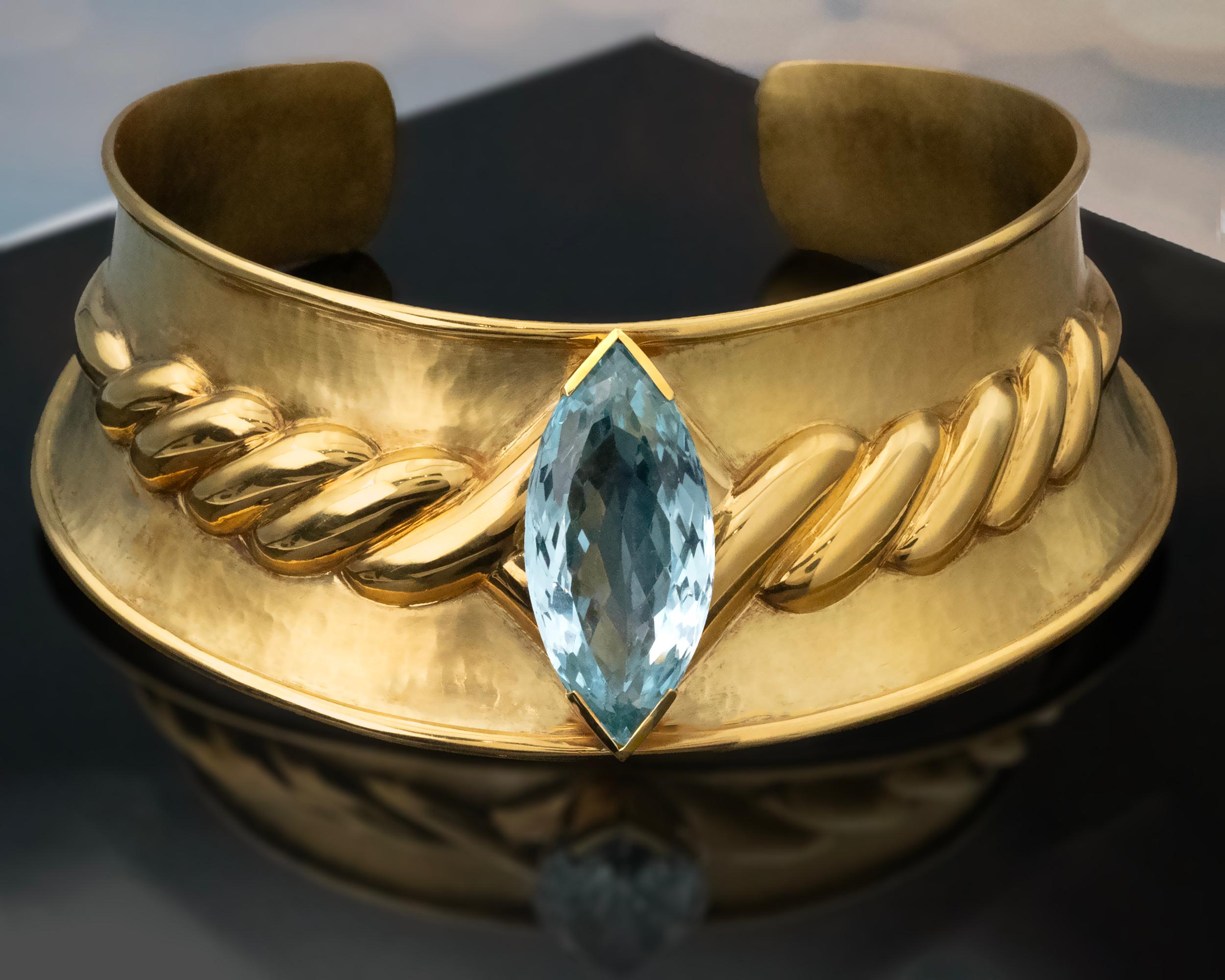 This out-of-the-ordinary 18K yellow gold cuff choker necklace showcases masterful craftsmanship, with a hand-hammered finish that enhances its unique character and elegance. At its center is a striking marquise-cut natural blue topaz, measuring