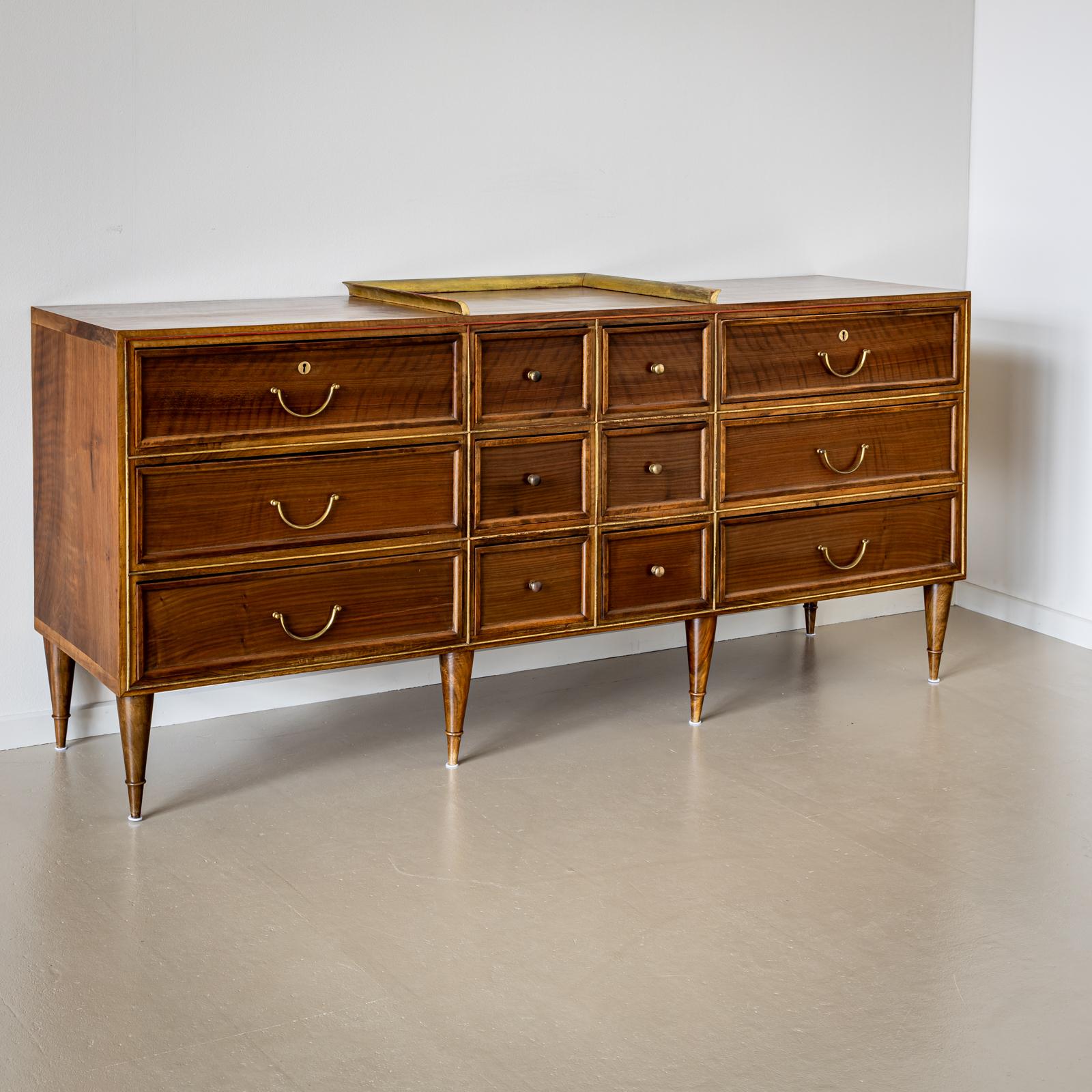 A mid century wide walnut veneered chest of drawers on six tapered legs with brass profiles and coffered drawer fronts. The chest of drawers is divided in the middle by a small U-shaped, gold-patinated gallery. The fittings are made of brass. The