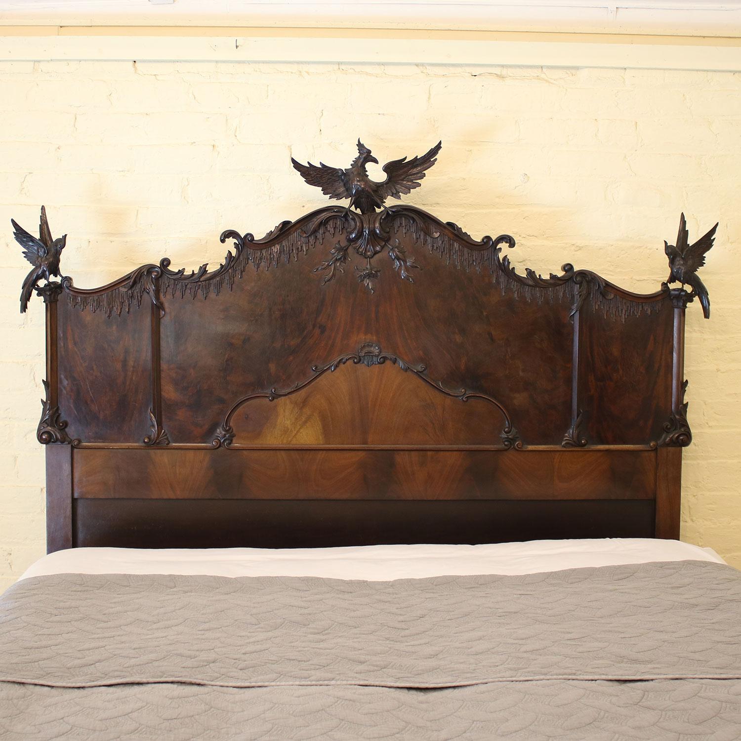 A Chippendale Revival bed from the early 20th century. The tall headboard is split into three panels of dark flame mahogany veneer and has a shaped top with scroll designs, crowned by three ornately carved birds of prey, possibly mythical eagles.