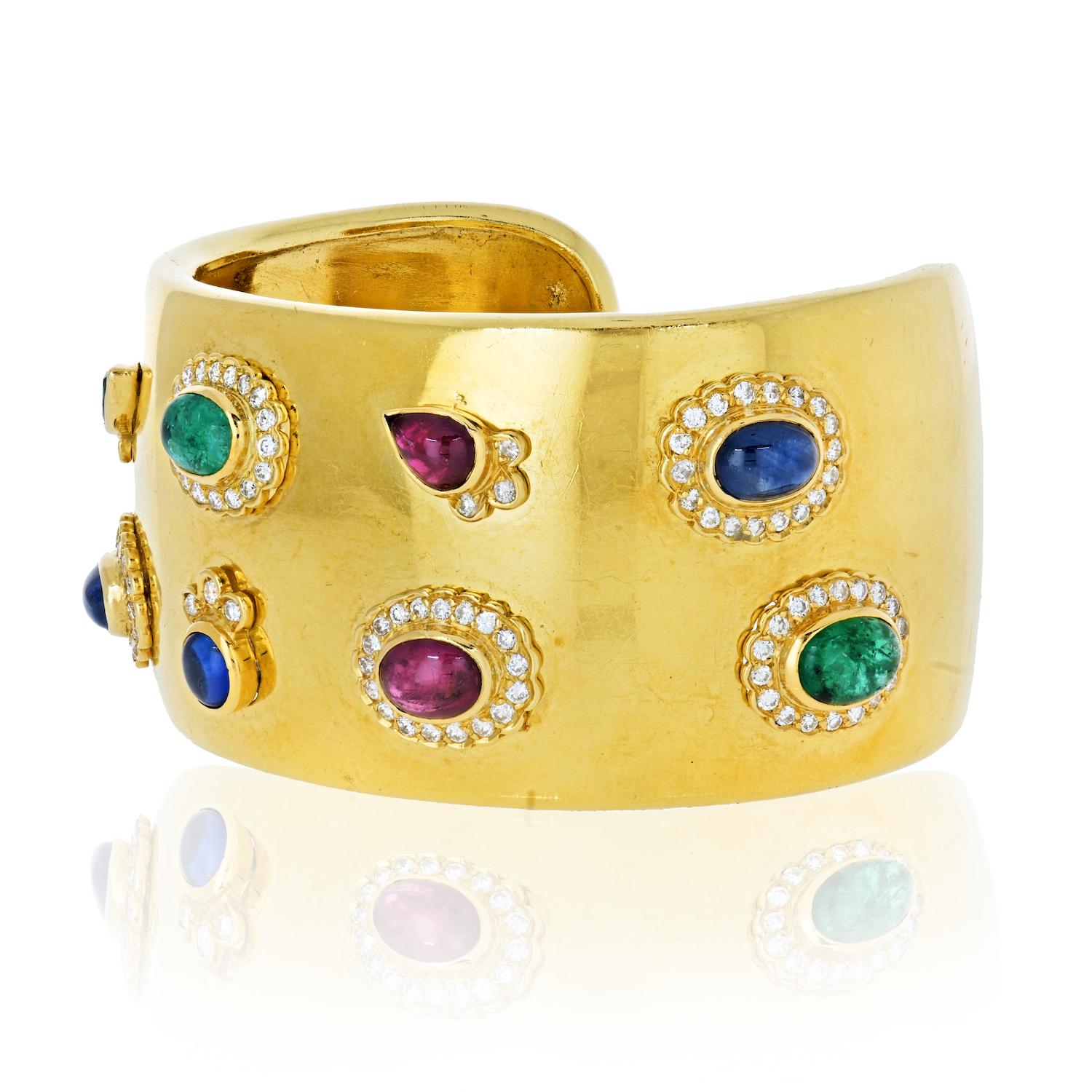 This is a vintage estate cuff bracelet at a statement-making size with colorful detailsin in 18k yellow gold with multicolored gemstones, including rubies, sapphires and green emeralds, and diamonds. Each gemstone is bezel set with a halo of mini