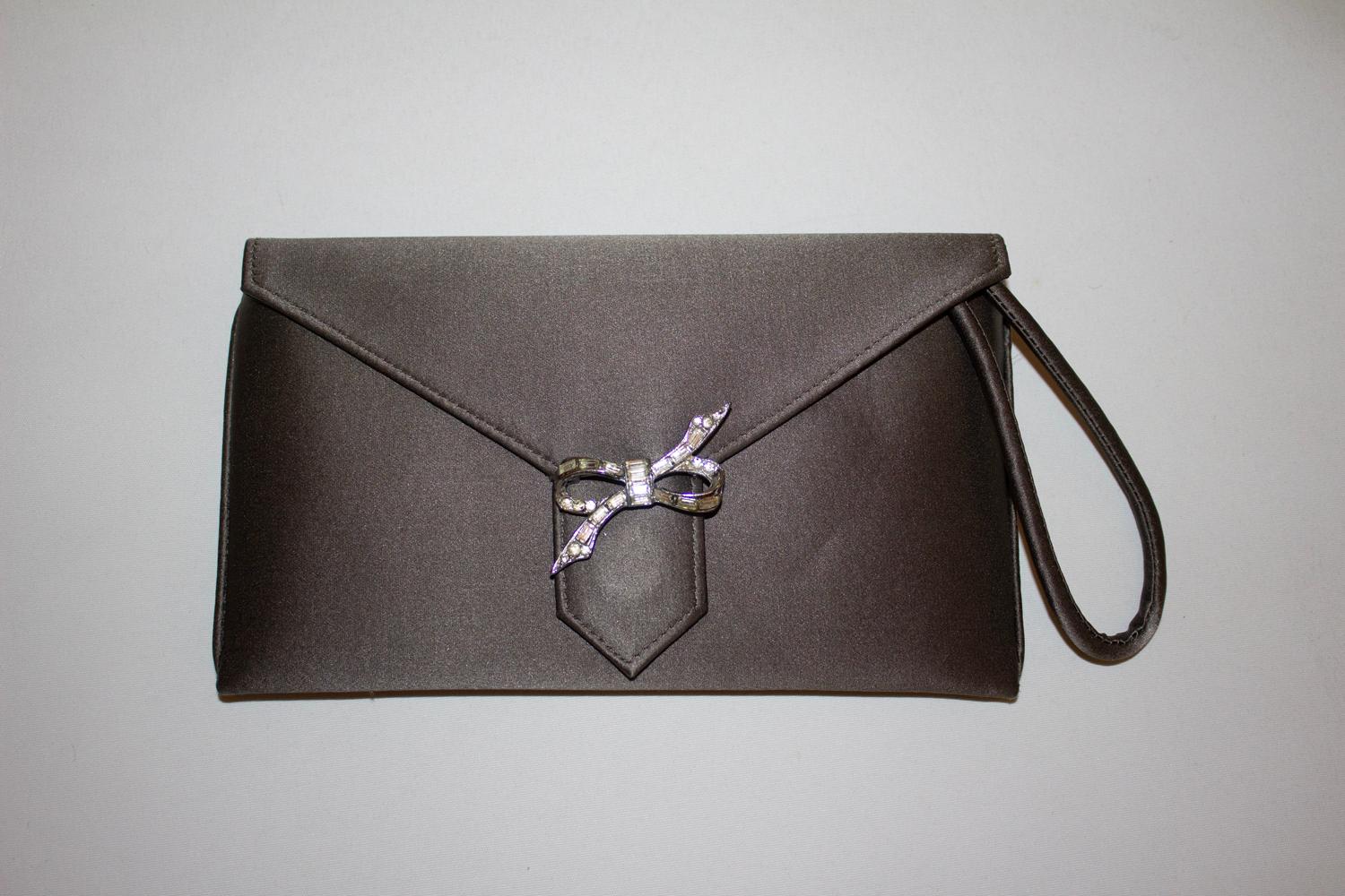 Eine einfach schicke Abendtasche von Wilbur und Gussie. Die Tasche aus attraktiver grauer Seide hat eine umklappbare Vorderseite mit einer hübschen Diamantenschleife.
Die  Die Tasche ist mit einer Beuteltasche und einem Handgelenksbügel ausgestattet
