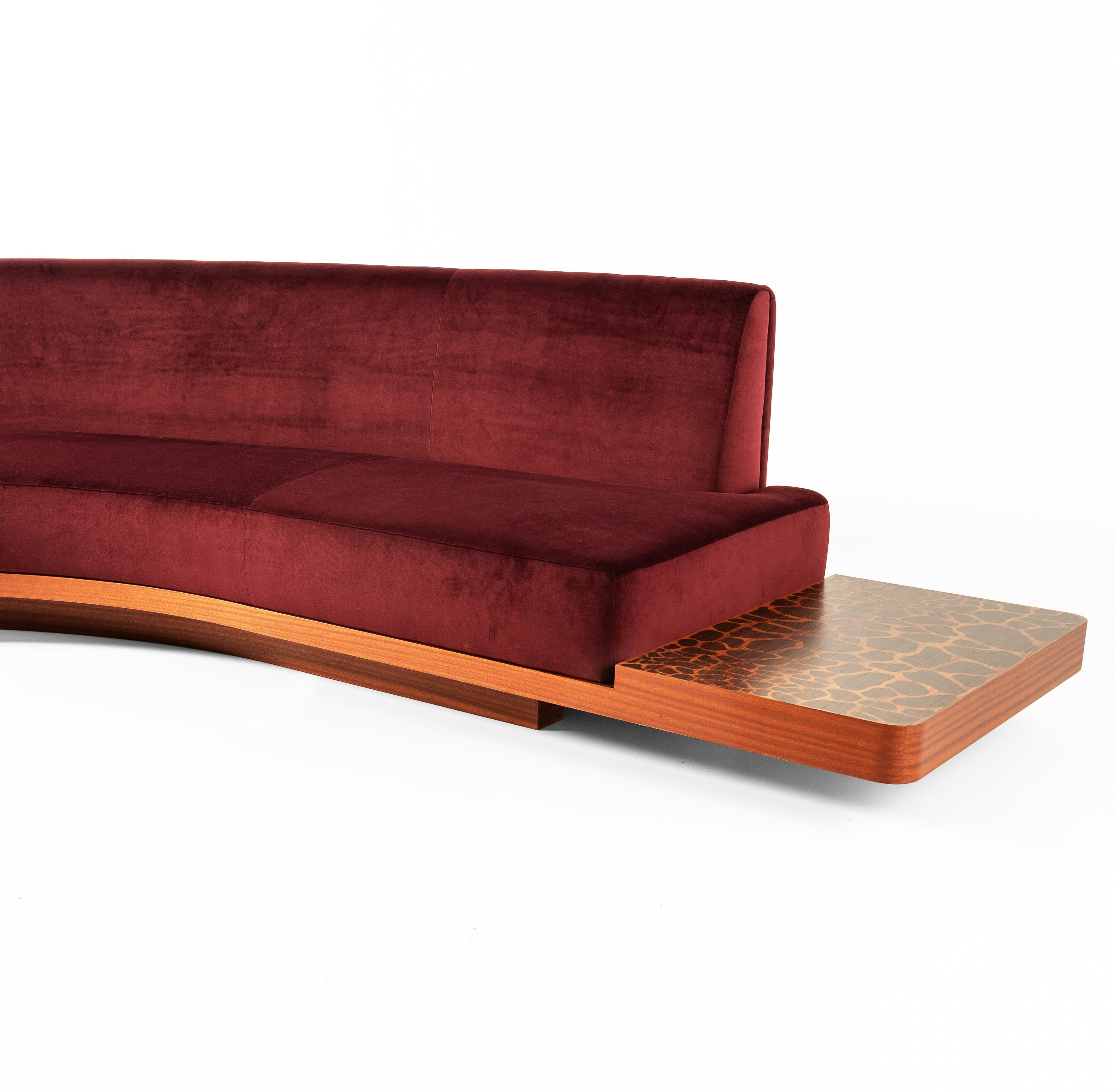 Wild Side Curved Red Wine Sofa with Table by Lorenza Bozzoli features a gently curved design upholstered in soft velvet with a deep red wine tone. The solid mahogany base provides stability and extends into a built-in low table with an eye-catching