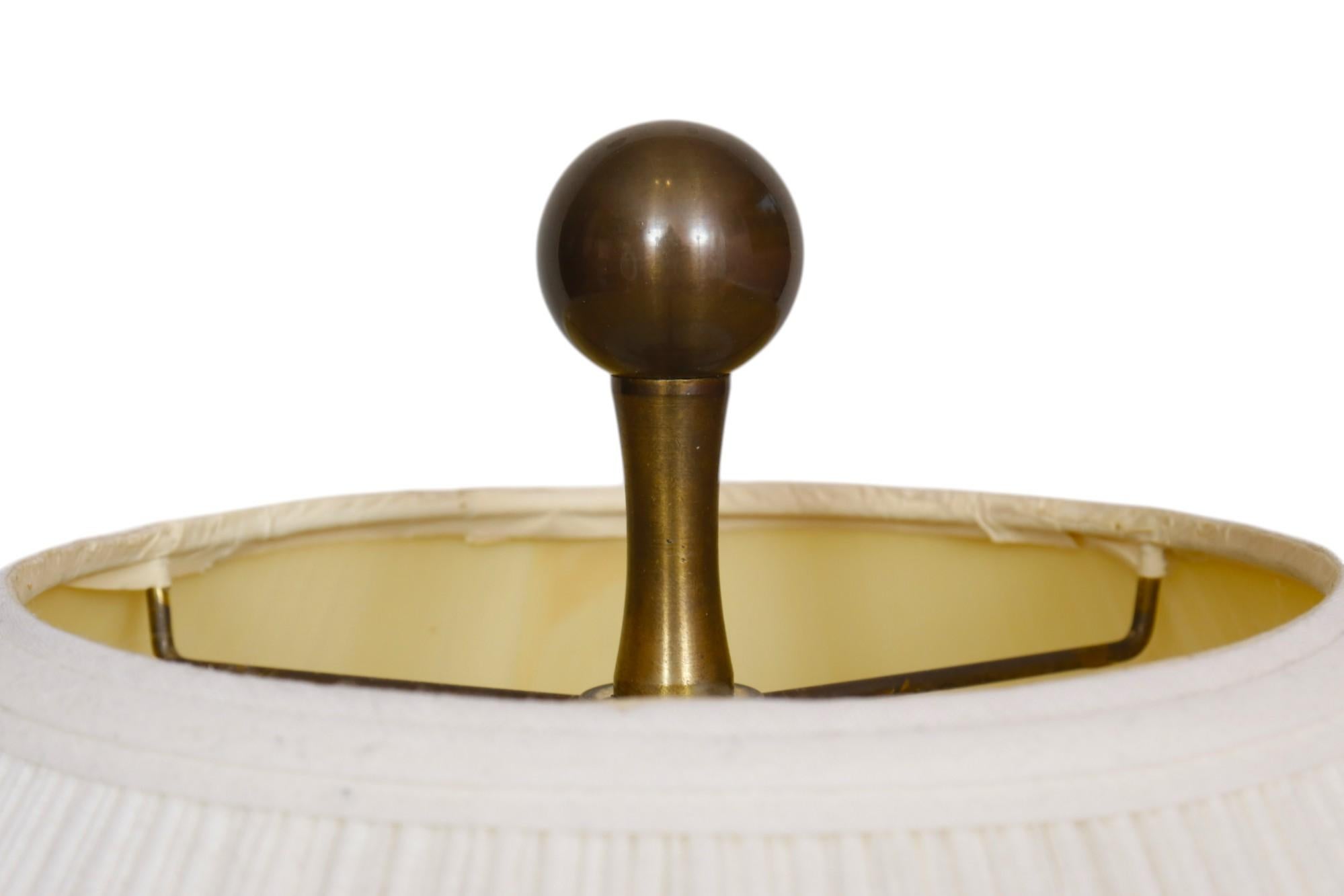 An amphora shaped brass table lamp made by Wildwood. A tall brass ball finial secures a rouched shade. The wide brass vase has two slender arms from the neck to the shoulder and sits on a formed resin base with Ming inspired feet. Measures 11