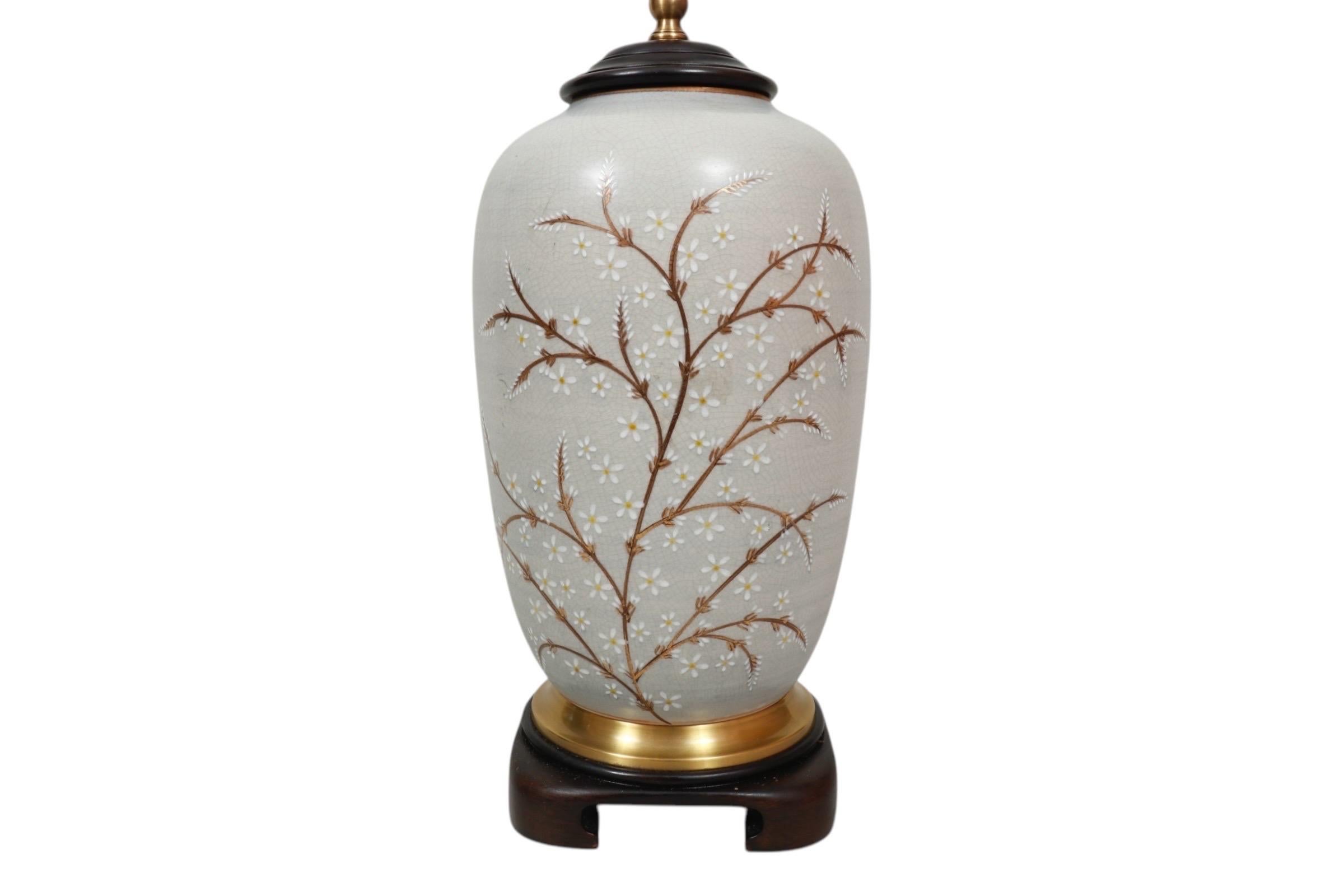 A Ming style table lamp made by Wildwood. A ceramic vase is hand painted with gilt foliage, dotted with white flowers, against a smokey taupe background. It sits in a brass ring above a wooden base carved with Ming feet. Above the vase is a beveled