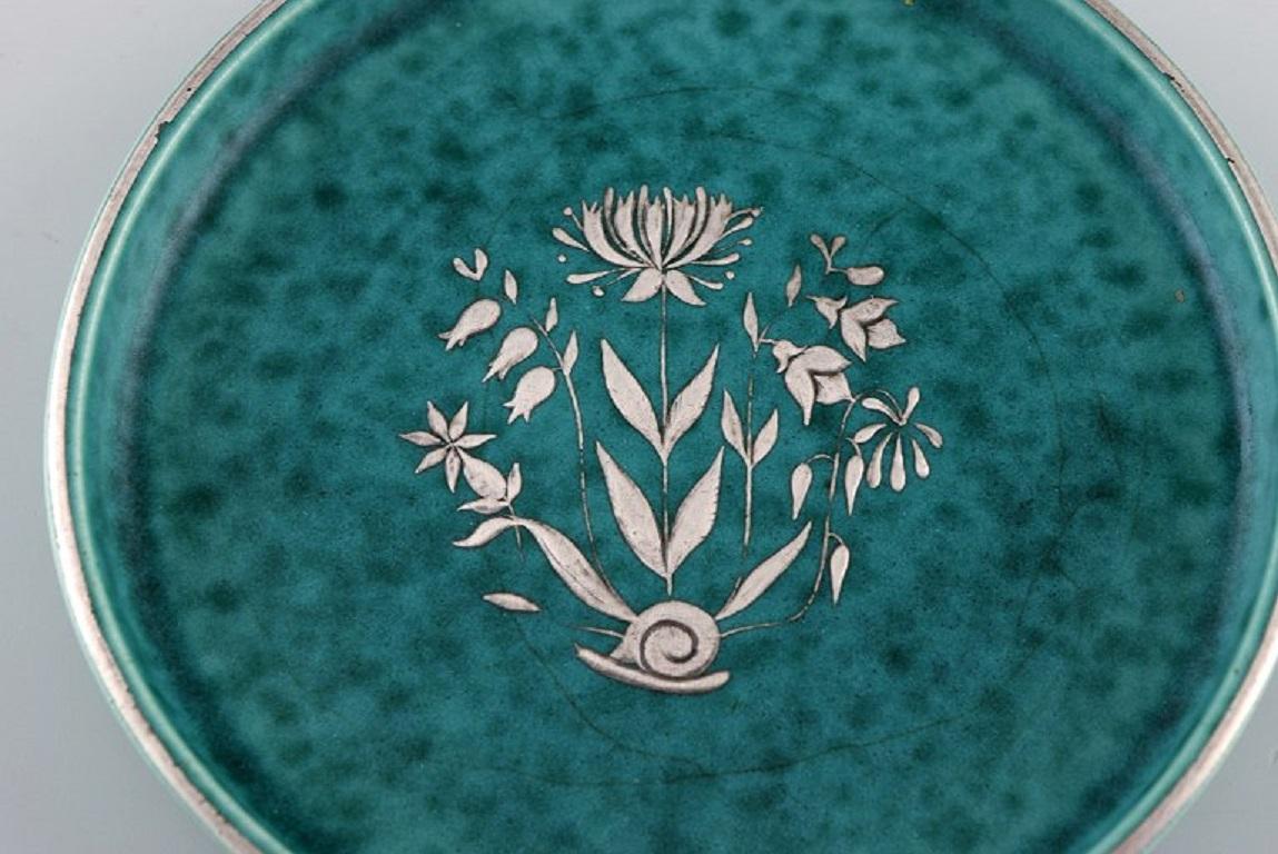 Wilhelm Kåge (1889-1960) for Gustavsberg. 
Round Argenta Art Deco dish in glazed ceramics. 
Beautiful glaze in shades of green with silver inlay in the shape of a flower. 
Dated 1938.
Measures: 13.5 x 2 cm.
In excellent condition.
Stamped.