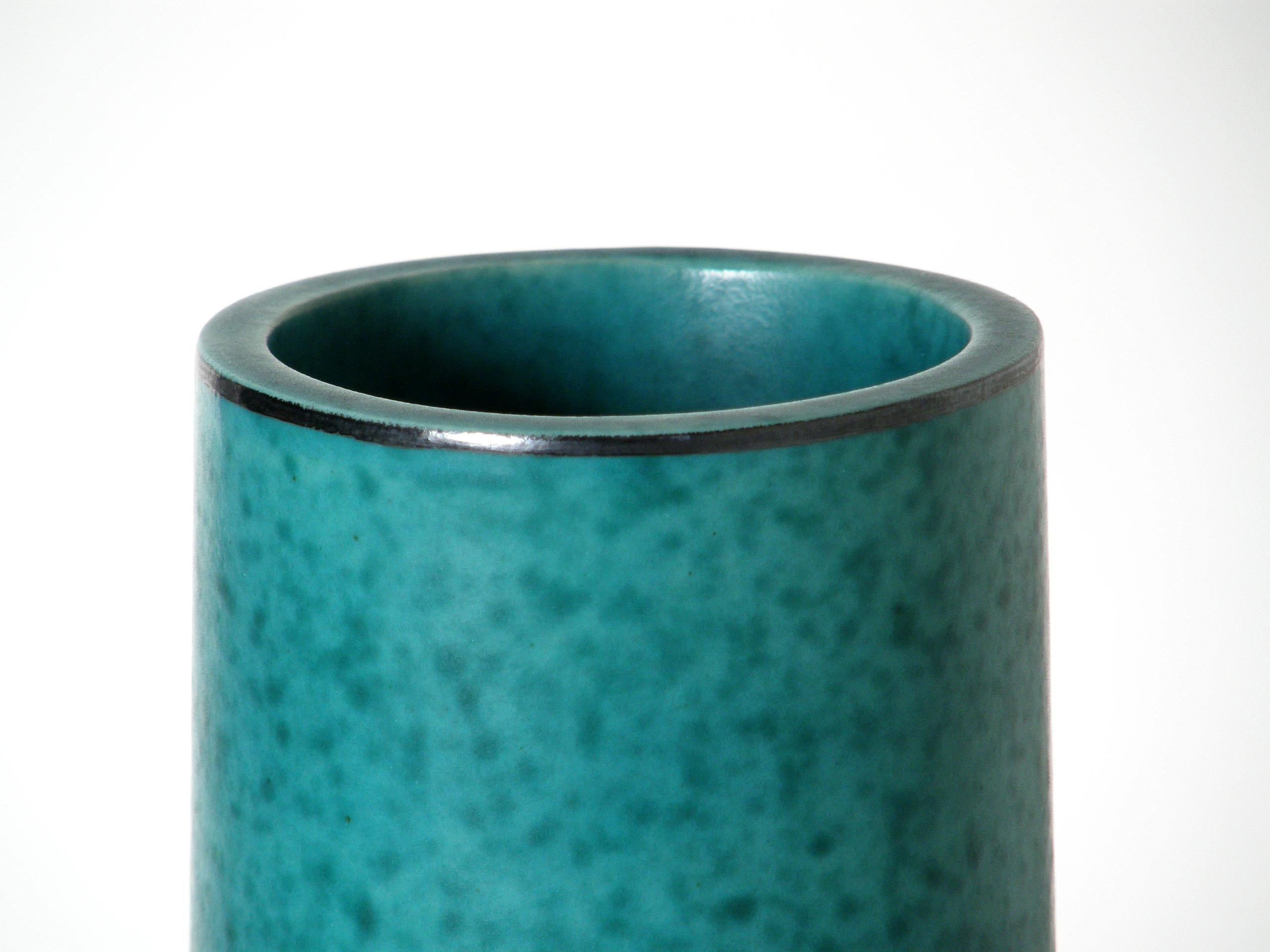 This classically elegant, columnar form ceramic vase is from Wilhelm Kåge's Argenta line for Gustavsberg. The mottled green glaze has a satiny finish, a floral silver overlay on the center front, and silver linear accents on the rim and foot.

We