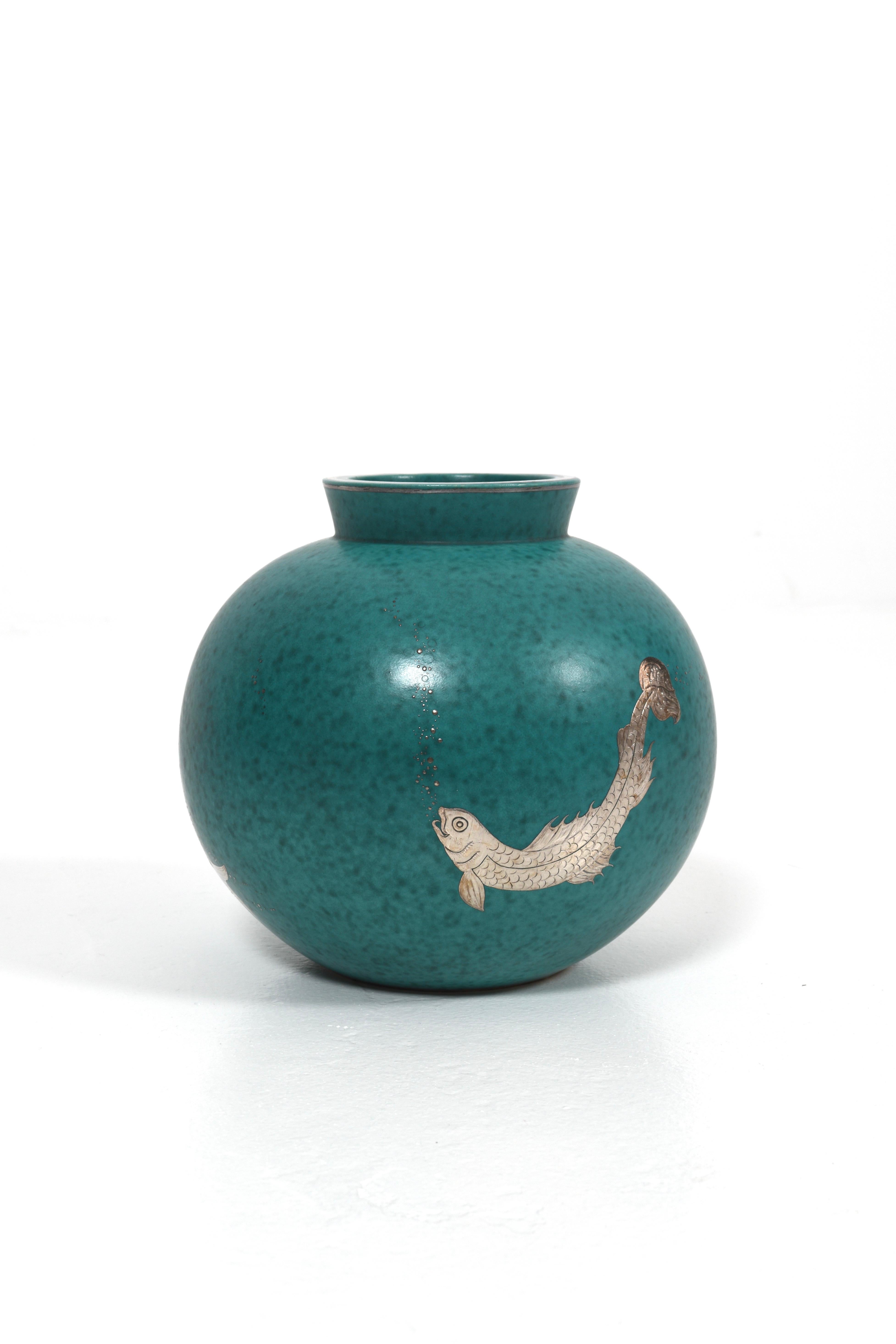 Este jarrón redondo de cerámica de la serie Argenta fue diseñado por Wilhelm Kåge para Gustavsberg en la década de 1940. Presenta un esmalte verde intenso con un acabado moteado y  está decorada con tres motivos diferentes de peces en plata. Los