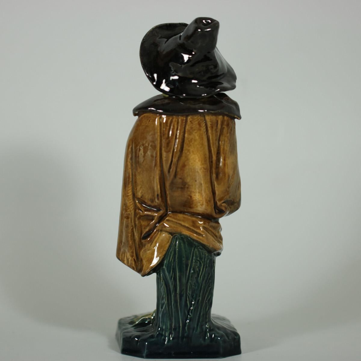 Schiller Majolica figure which features a frog playing bagpipes, dressed in a cloak and a hat. Coloration: green, light brown, yellow, are predominant. The piece bears maker's marks for the Schiller pottery. Bears a pattern number, '1107'.