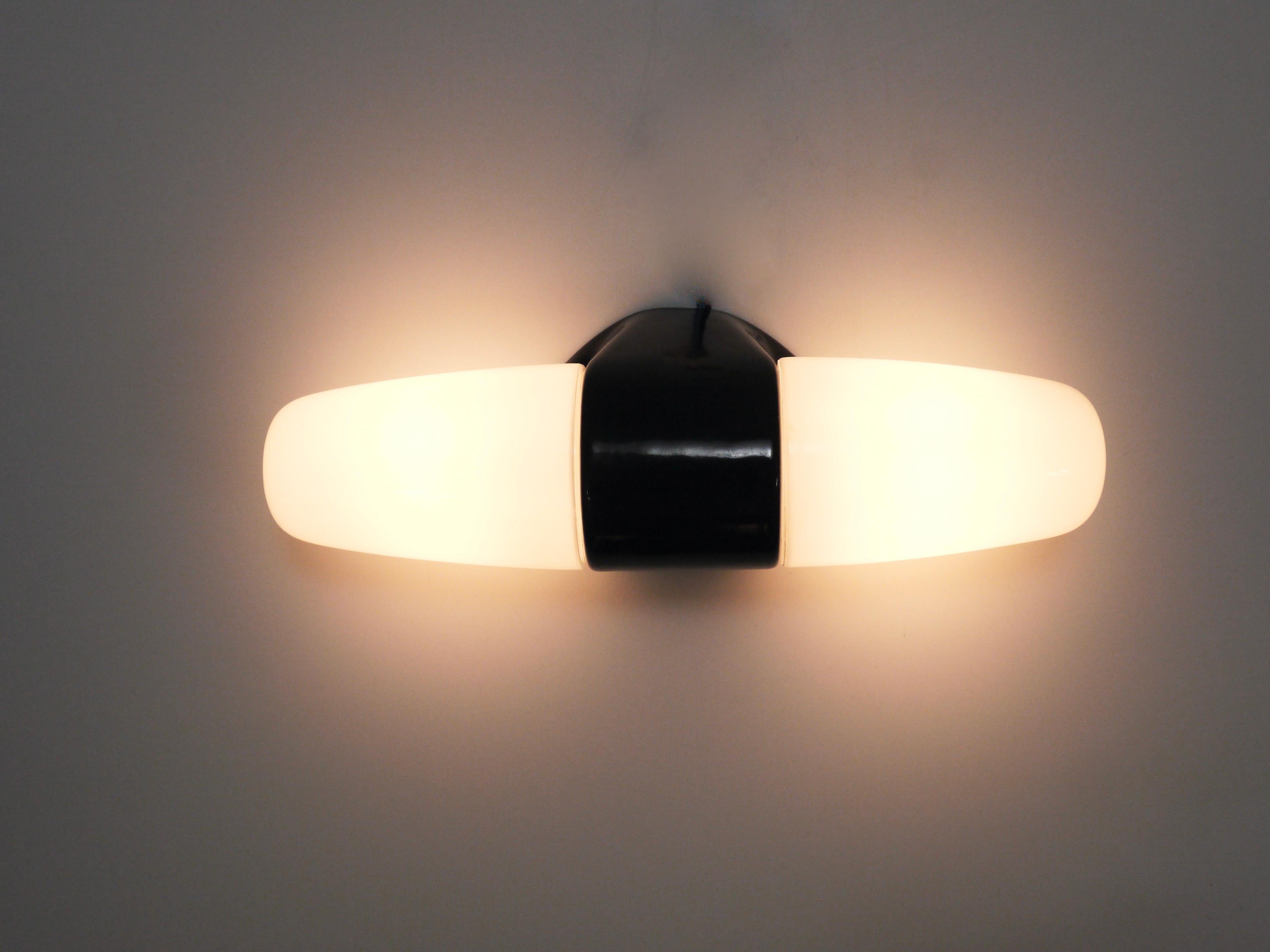 A Midcentury Modern double socket wall lamp/sconce dating back to the 1950s, designed by Wilhelm Wagenfeld. Made of porcelain with a glossy black glaze accentuated by two white opaline glass lampshades/bulb covers. Based on our experience so far,