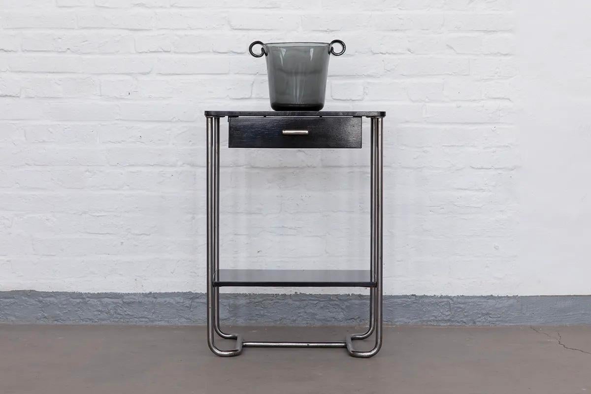 Dieser Weinkühler aus Rauchglas wurde 1937 von Wilhelm Wagenfeld entworfen und von den Lausitzer Glaswerken in Weißwasser, Deutschland, hergestellt.
Das aus Pressglas gefertigte Stück spiegelt mit seiner klaren Form und zurückhaltenden Eleganz den