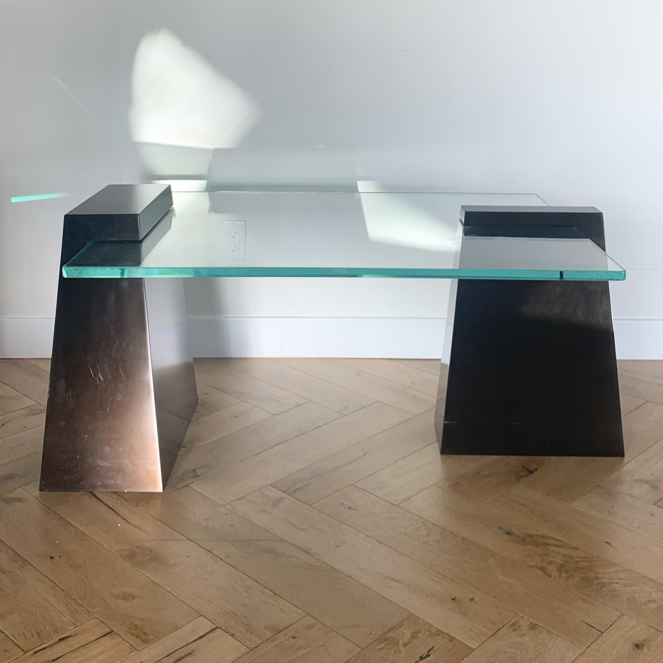 A vintage Will Stone or Karl Springer style modernist coffee table featuring two metal asymmetrical plinths that swallow a thick glass slab, late 20th century. The plinths are a sumptuous bronze, recalling postmodern industrialism. One is even