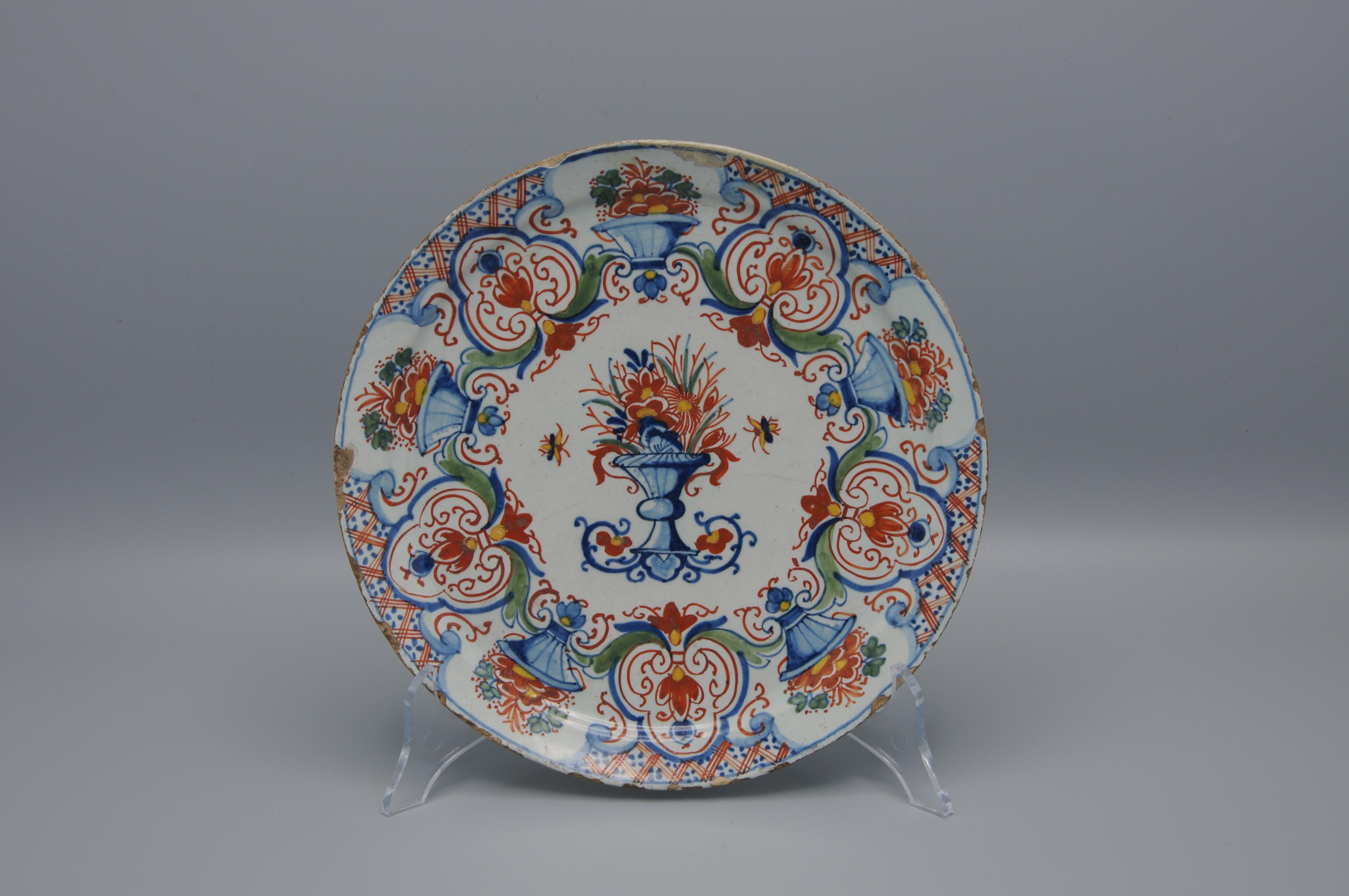 Piatto policromo olandese di Delft con vivace decorazione policroma di un vaso centrale su un supporto pieno di fiori affiancato da due piccoli insetti. Bordo molto decorato con cartigli alternati con vasi di fiori simili e volute fogliate.

Mark WK