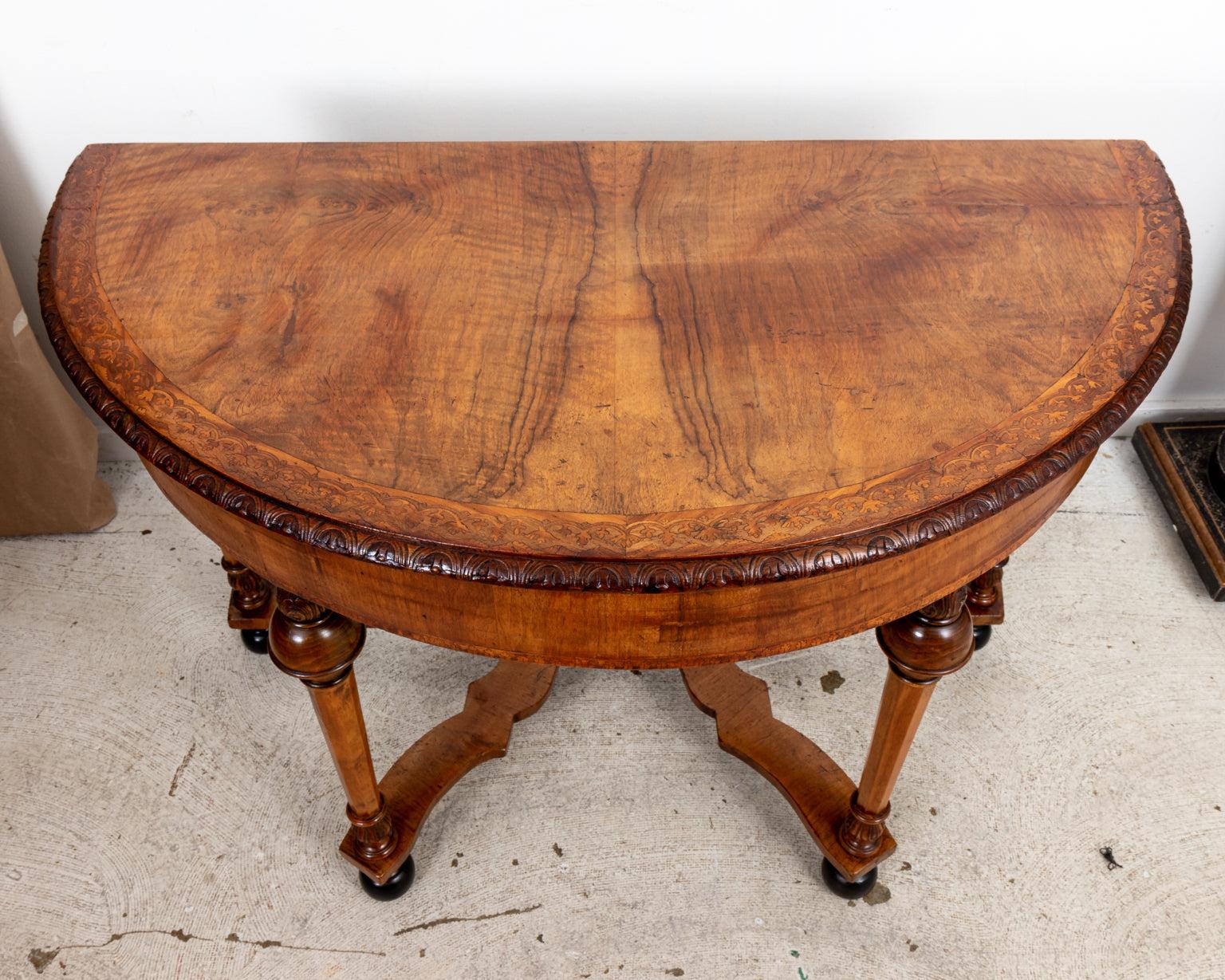 Circa 1900s William & Mary style Walnut demilune table with carved wood detailing and inlay work throughout. The skirt is detailed with carved foliage trim while the edge of the tabletop is crafted with foliage inlay in a similar pattern. The base