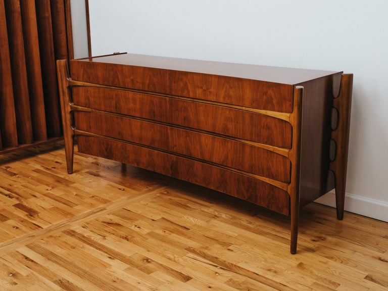 A large chest of drawers or credenza, its case with a concave façade in bookmatched walnut and suspended within an external frame of vertebral legs. Drawers have concealed pulls and beech interiors with partitions to the top pair. Heavy, robust