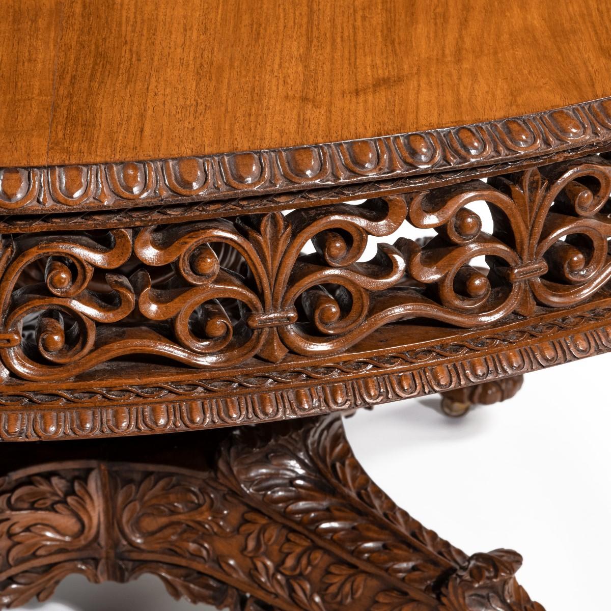 A William IV Colonial padouk five-foot round table, the solid circular top with a pierced frieze of paired volute scrolls, with blind fretwork and egg-and-dart borders, the baluster support with deeply carved leaves, the four S-scroll legs further