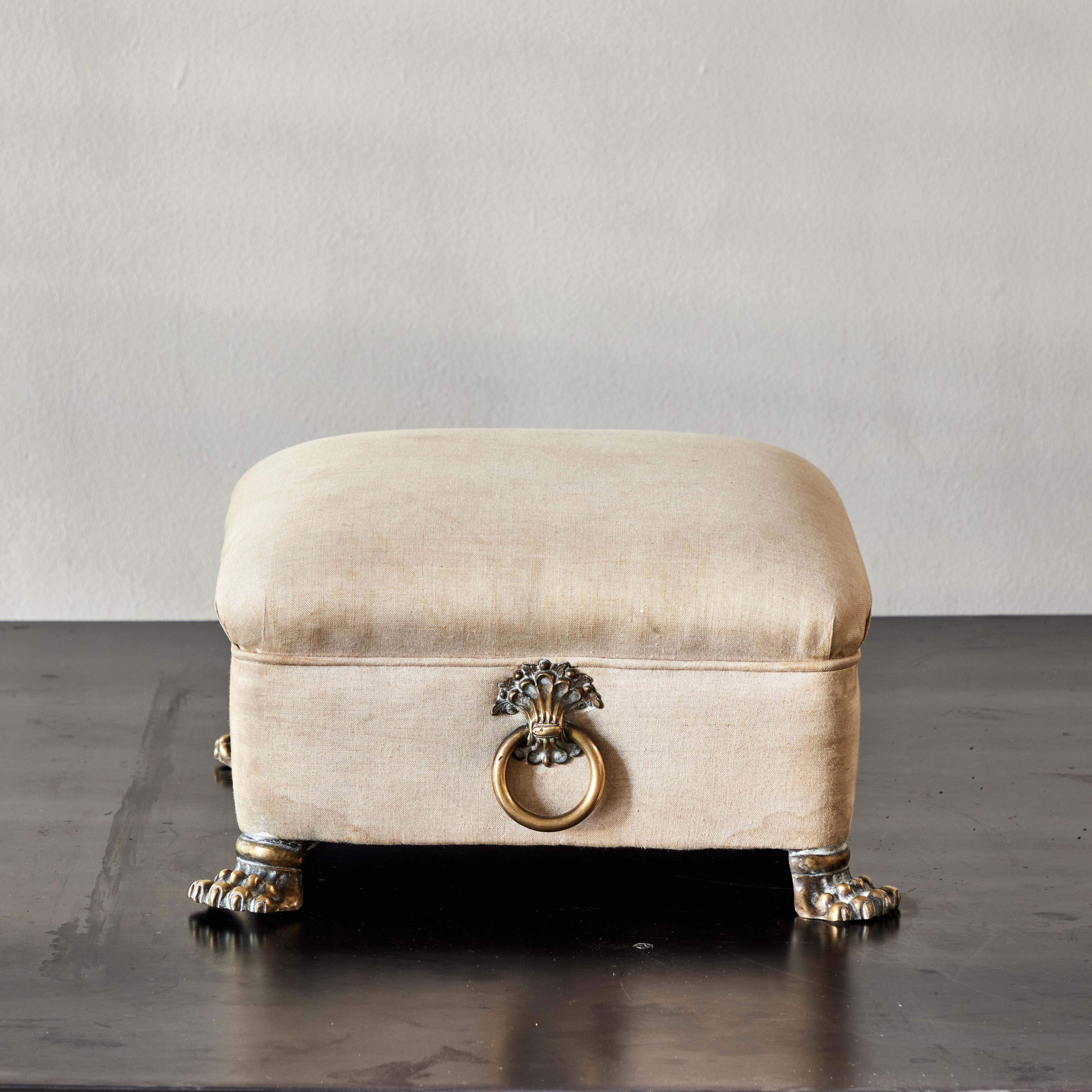 William IV era footstool with decorative brass claw feet and handle rings, upholstered in cream-colored linen. A uniquely charming addition to any seating arrangement. 

England, circa 1830

Dimensions: 14W x 14D x 8H.