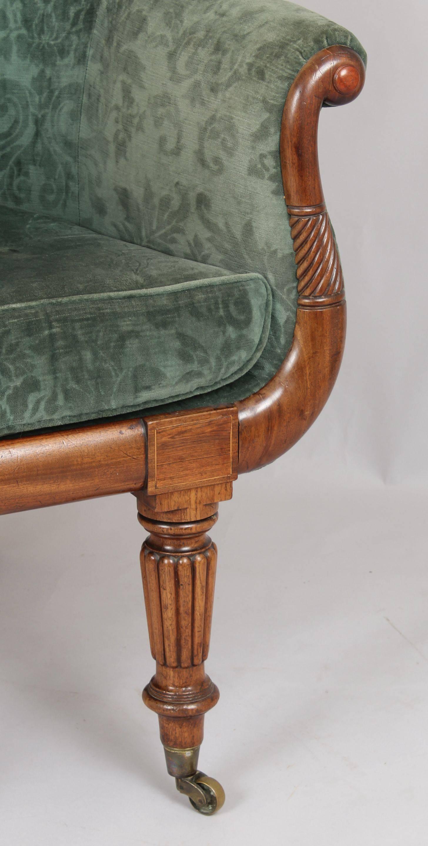 William IV period tall-backed easy-chair; the fully-upholstered curved back and squab seat covered in dark green patterned velvet; with scrolled and spiralled mahogany arm-facings and bold turned and reeded fore-legs with brass cup-castors.