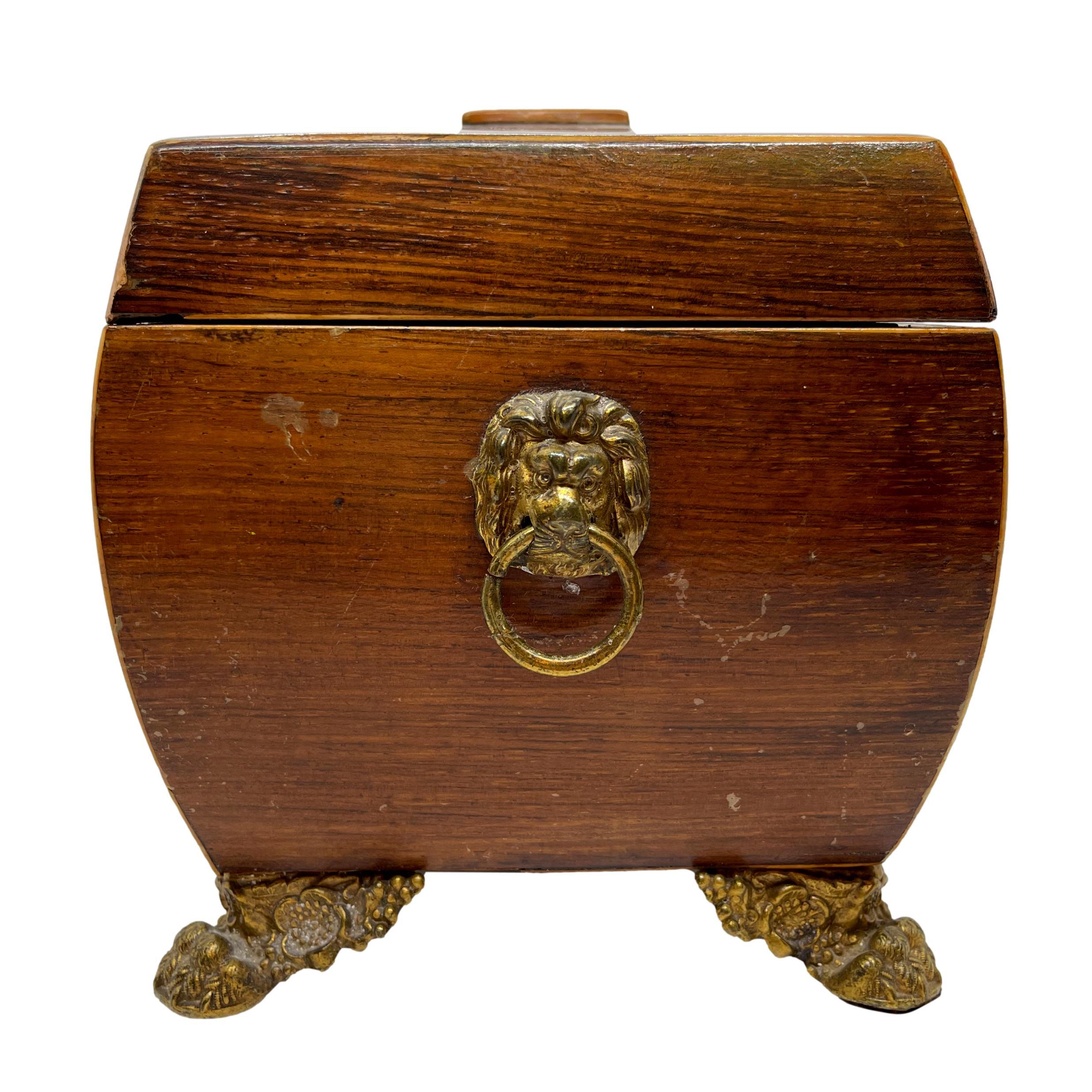 William IV rosewood and satinwood-banded tea caddy, English, ca. 1835, of sarcophagus shape, with lion mask ring pulls to either side, the fitted interior with two lidded tea compartments and space for the mixing bowl (not present), on molded brass