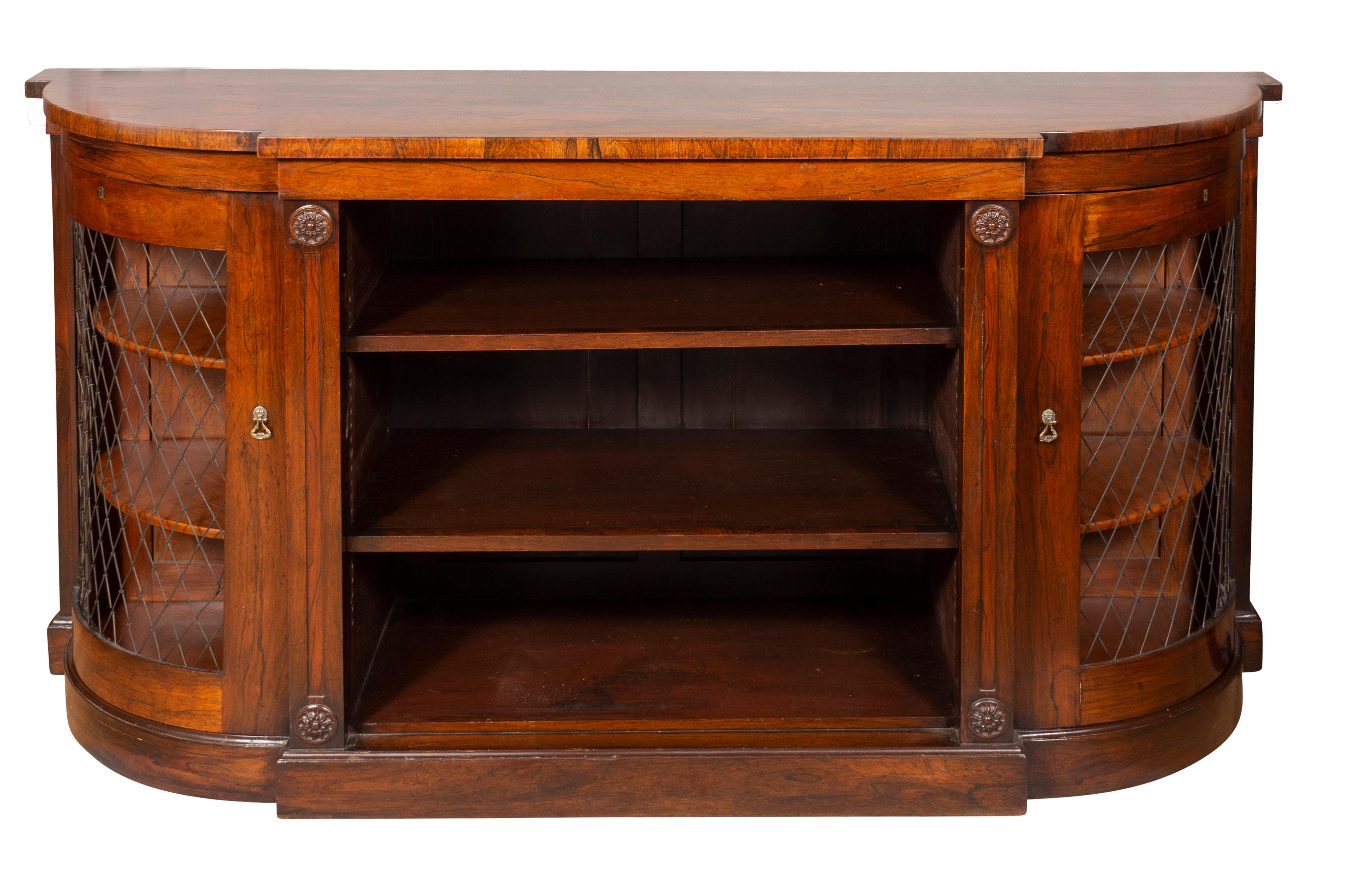 With rectangular top with rounded ends over a conforming case with central open bookcase containing two adjustable shelves, flanked by a pair of curved grill cabinet doors. The case with circular carved rosettes. Plinth base.