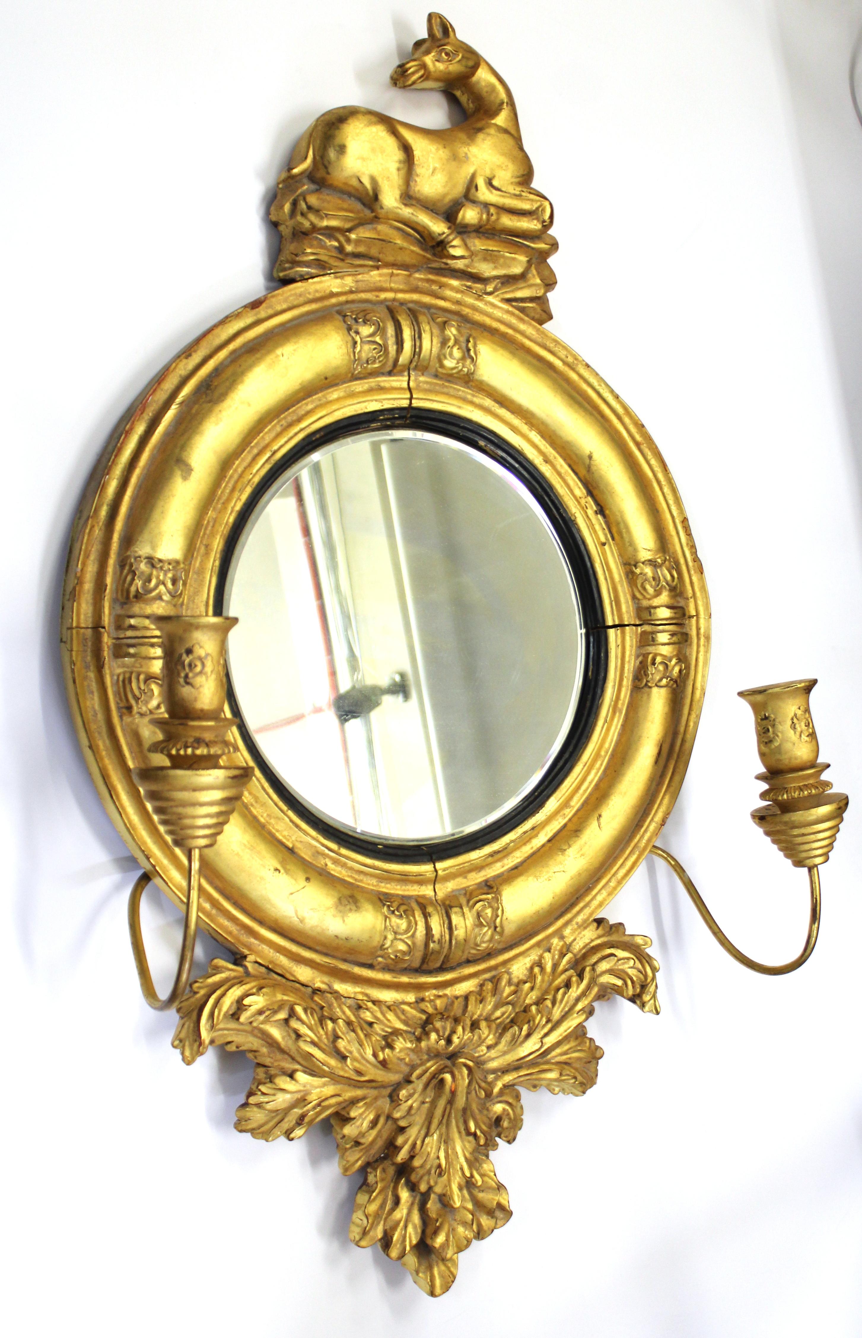 William IV style round girandole mirror made in giltwood, with decorative foliage and a resting heraldic animal surrounding a circular flat mirror with a half-rounded frame with carved lotus leaf clasps. Two candle-holding arms, one on each side of