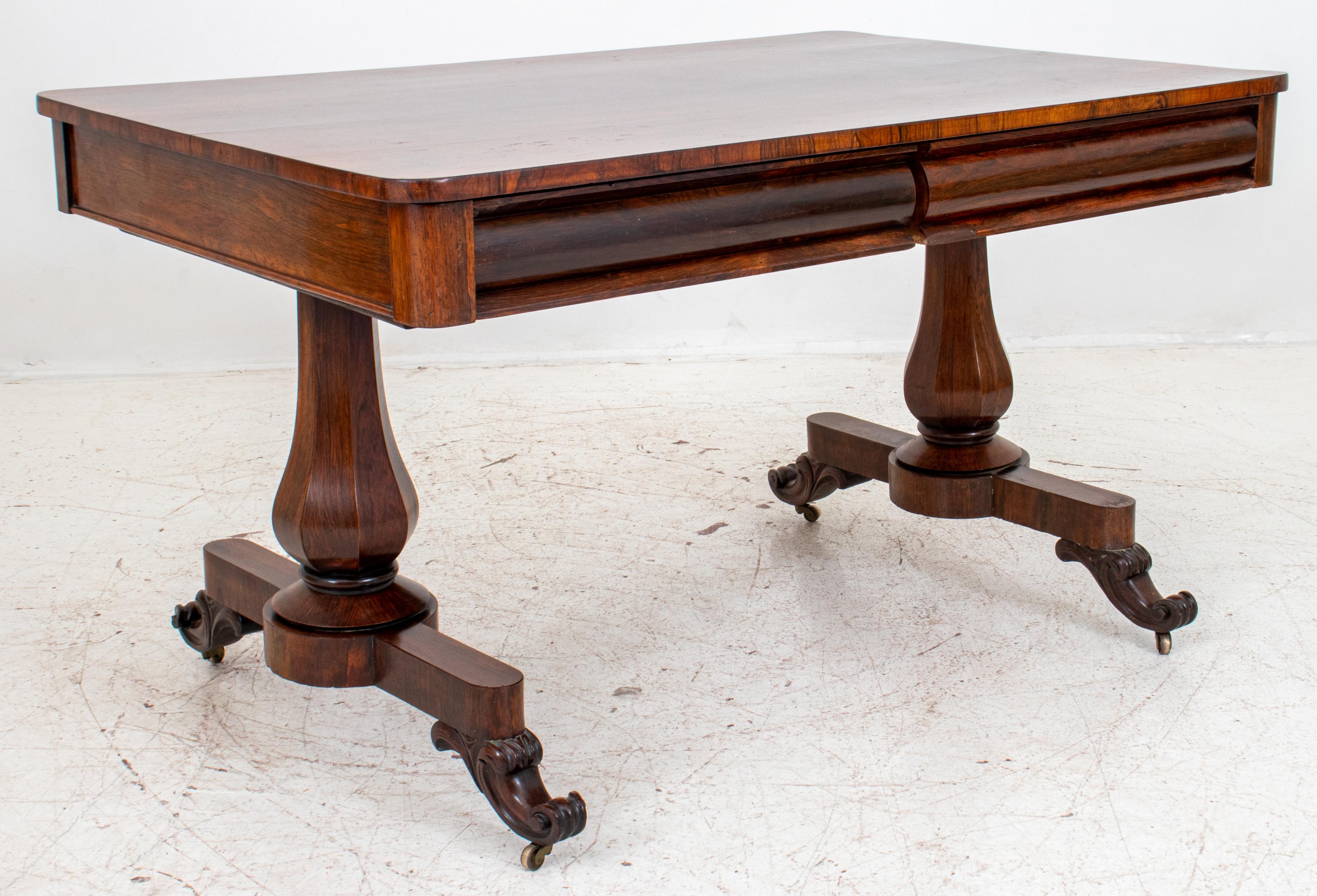 William IV style rosewood library table or partners desk, rectangular, the apron concealing four drawers behind convex rounds on two hexagonal baluster supports on splayed legs terminating in brass casters. Measures: 29