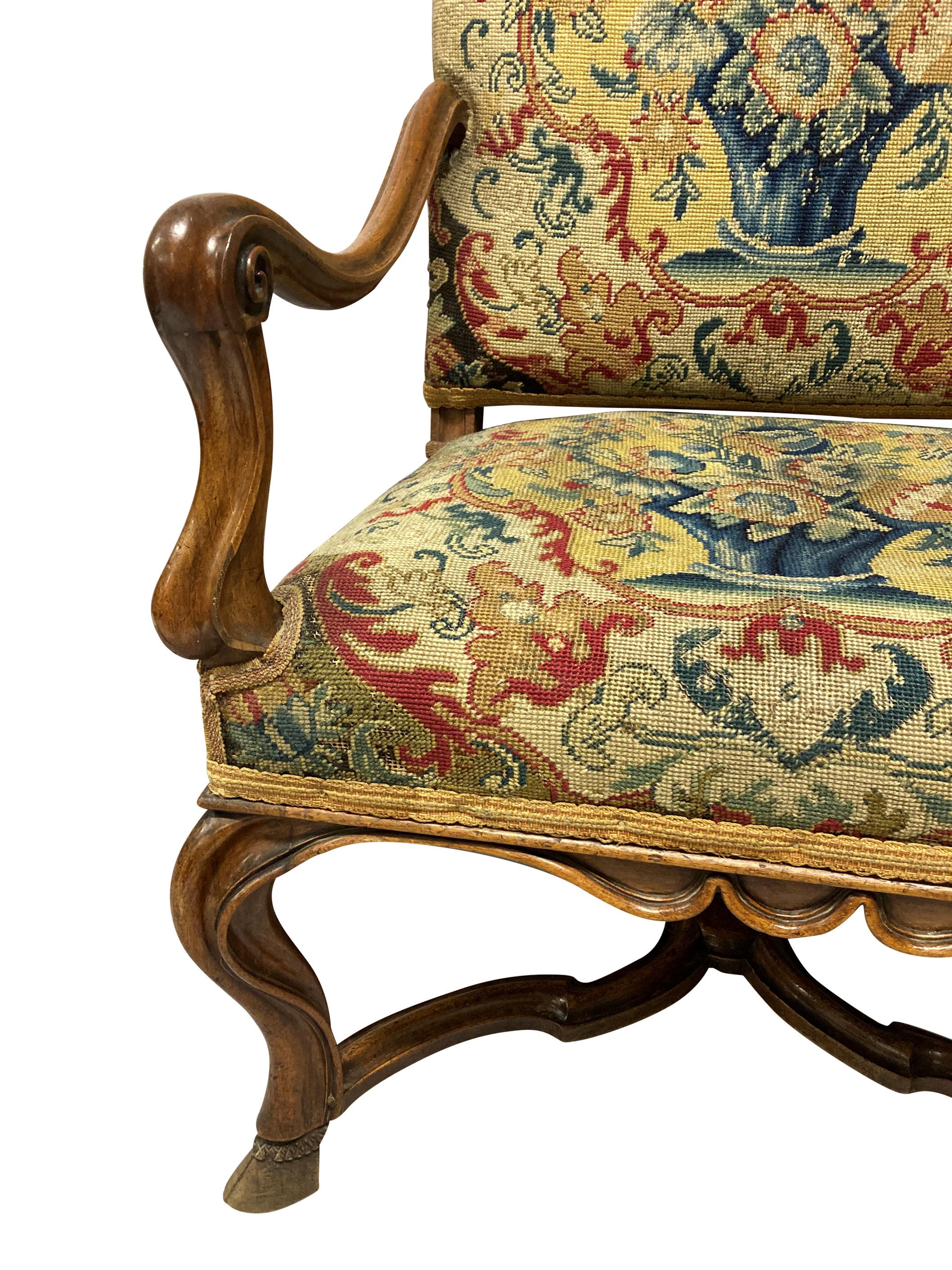 A fine William & Mary carved walnut armchair of good colour, with scrolled arms, stretcher and deer feet (the back two have partly worn down). Upholstered in an early XIX Century needlepoint tapestry in good condition.