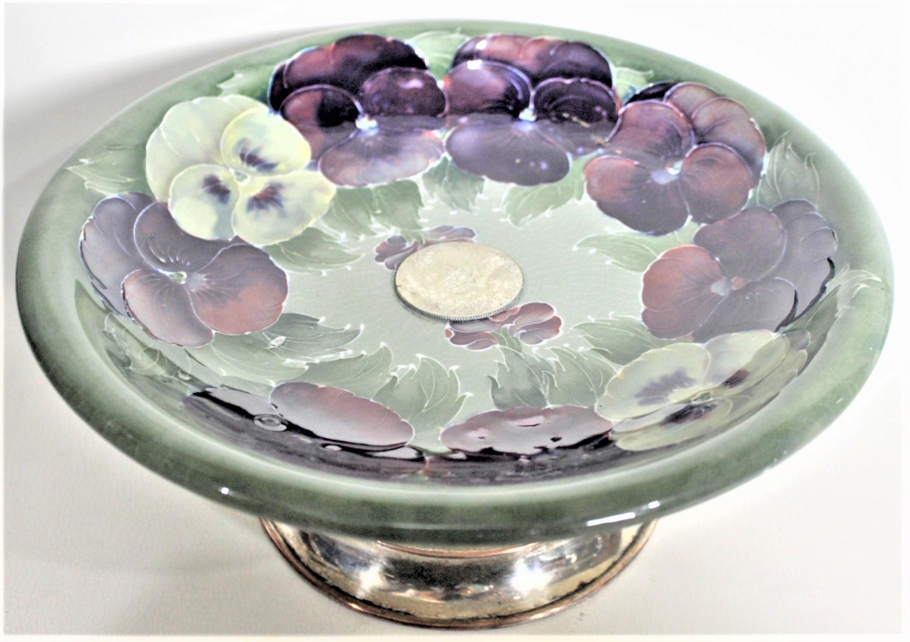 This art pottery pedestal bowl or dish was done by the Moorcroft Pottery Company of England in circa 1925 in their early 'Pansy' pattern. The dish is done with a deep green ground with vibrant purple and cream colored pansies around the inner
