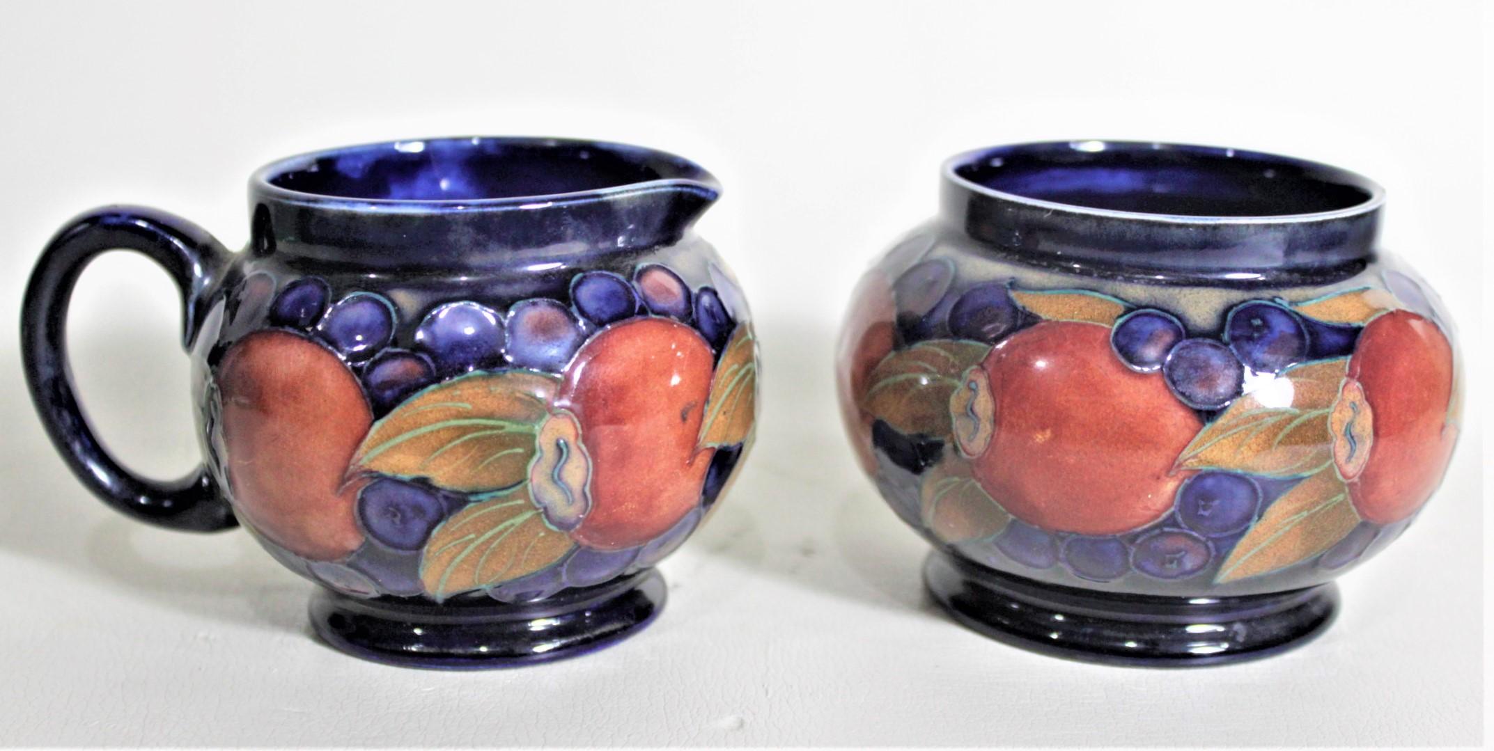 This art pottery cream and sugar bowl set was done by the Moorcroft Pottery company of England in circa 1925 in their 'Pomegranate' pattern. This duo is done in the Moorcroft signature deep cobalt blue ground with a series of fruit around the