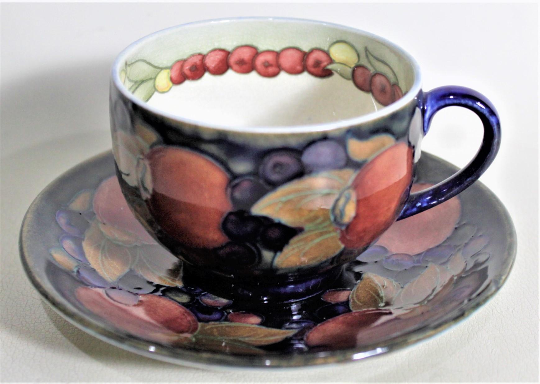 This art pottery teacup and saucer set was done by the Moorcroft Pottery company of England in circa 1925 in their 'Pomegranate' pattern. Each duo is done in the Moorcroft signature deep cobalt blue ground with a series of fruits around the outer