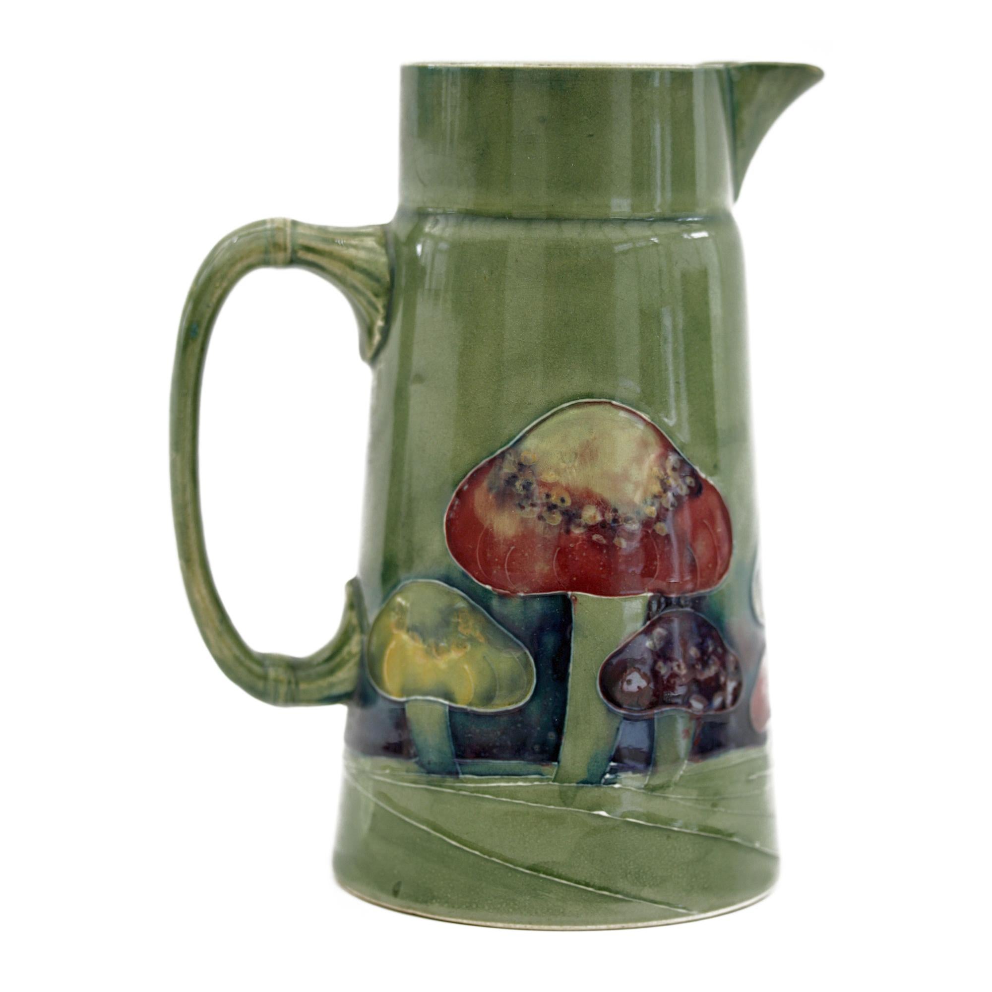 A rare William Moorcroft Claremont pattern jug of tall conical shape with a moulded handle and funnel shaped top with large pouring spout. The jug is hand decorated with tubelined mushrooms in streaked blue, red and yellow glazes on a green ground.