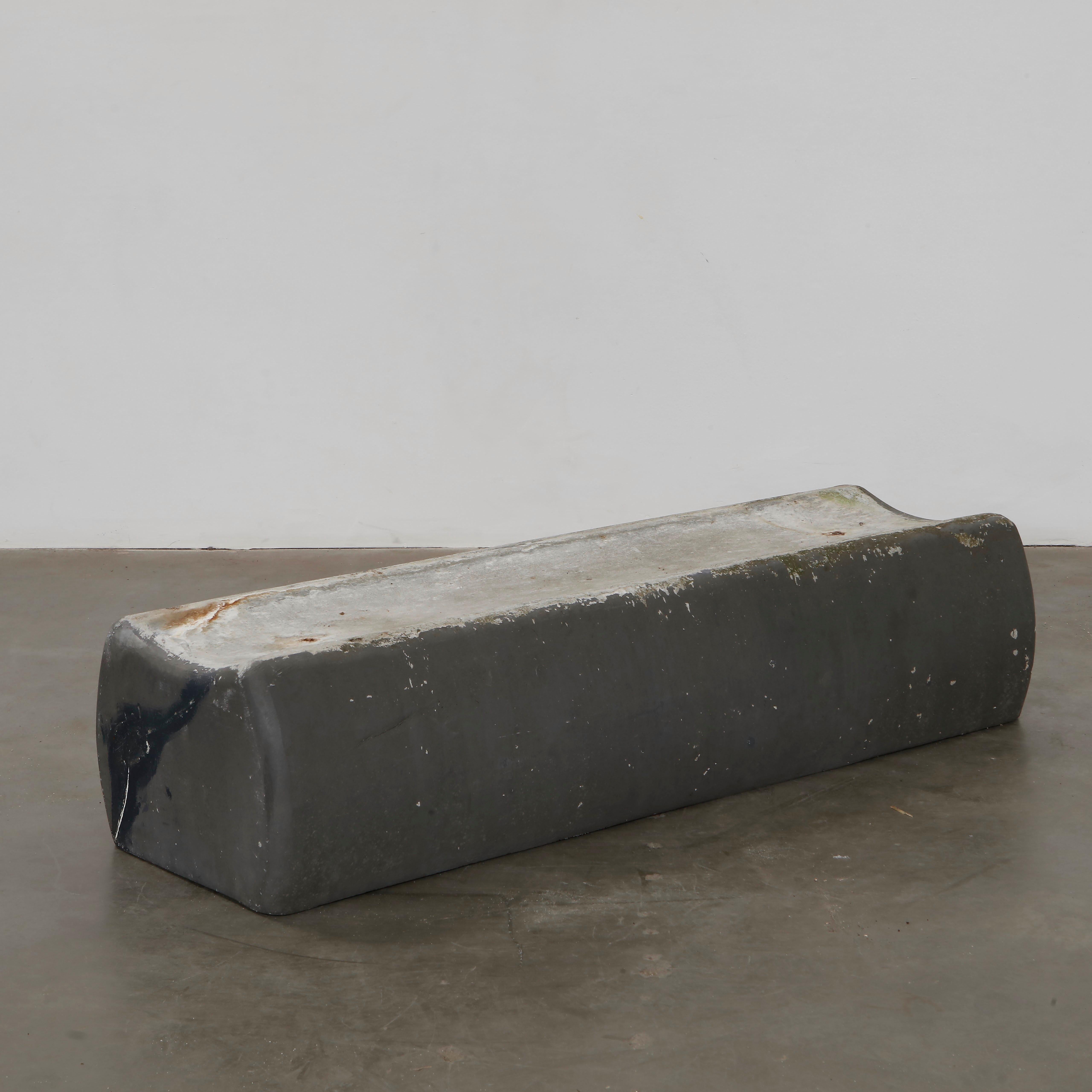 A heavy, minimalist concrete bench designed by Willy Guhl. Featuring a simple, curved structure, it embodies brutalist aesthetics. The raw texture and natural wear give it a striking sculptural quality. Height: 13 in. Width: 59 in. Depth: 17 in