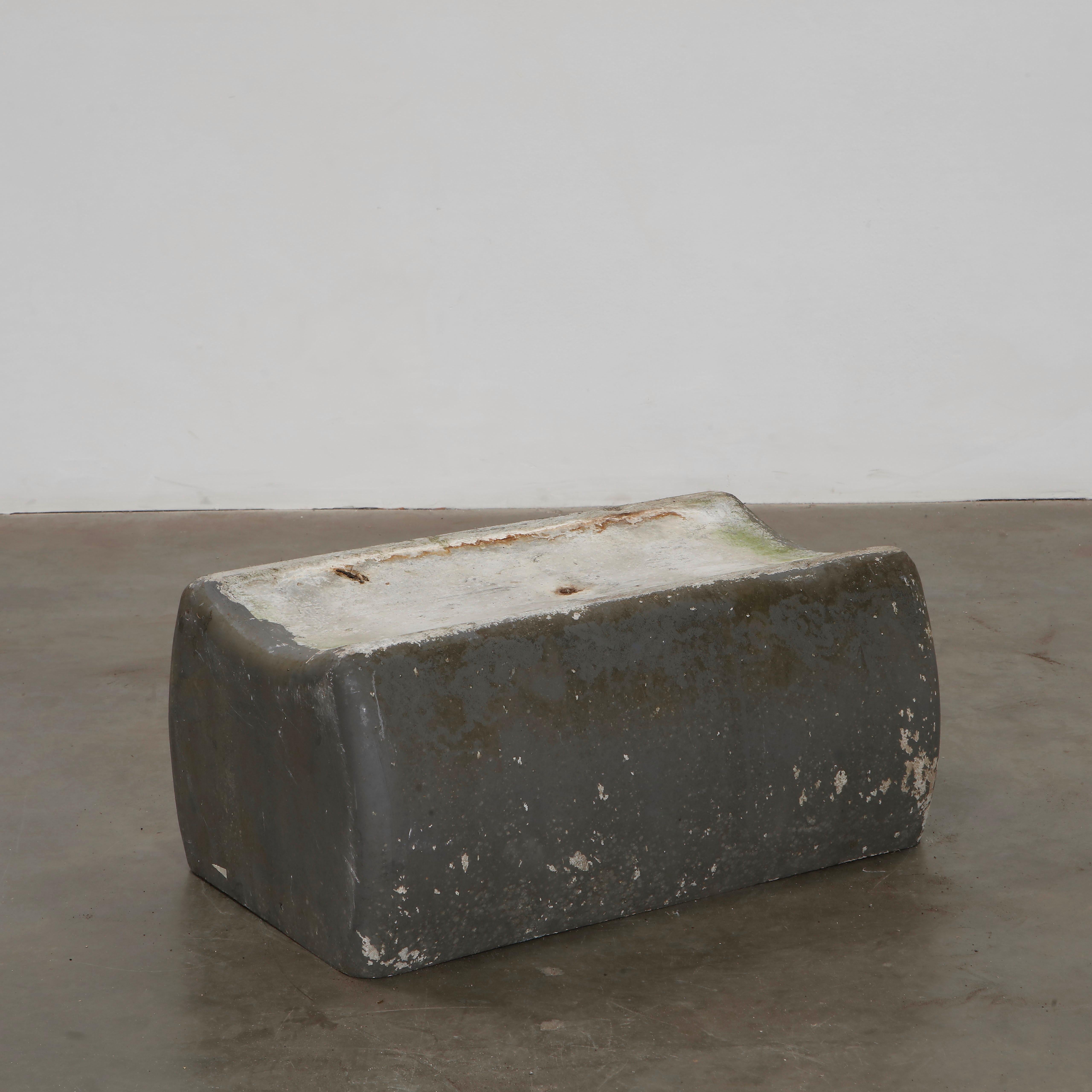 A heavy, minimalist concrete bench designed by Willy Guhl. Featuring a simple, curved structure, it embodies brutalist aesthetics. The raw texture and natural wear give it a striking sculptural quality. Height: 13 in. Width: 29.5 in. Depth: 17 in.