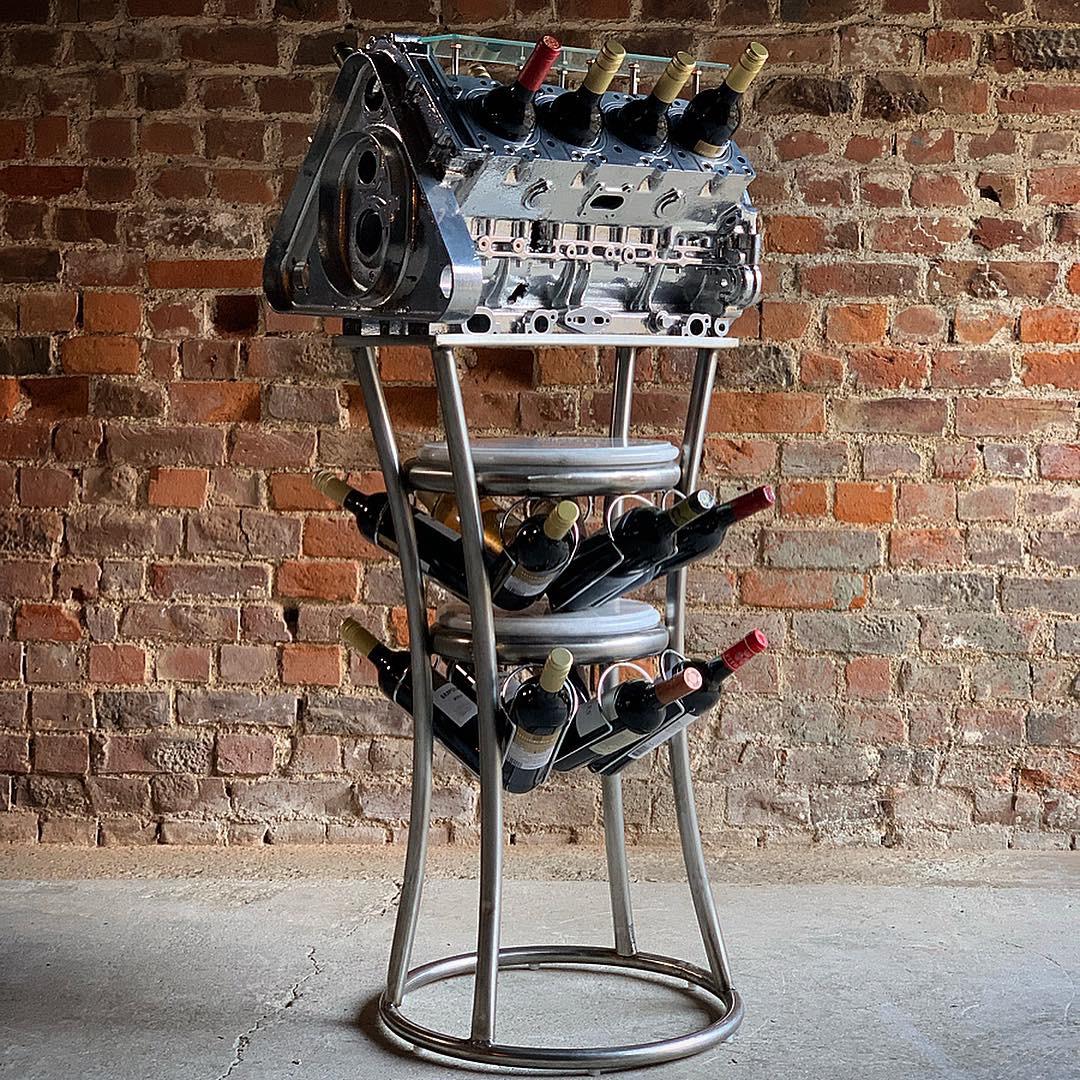 Stunning chrome coated V8 engine block converted to a twenty bottle wine rack, the chromed engine with frosted glass top with eight bottle holder piston apertures, raised on a tubular steel stand with two white marble tiers and an arrangement of