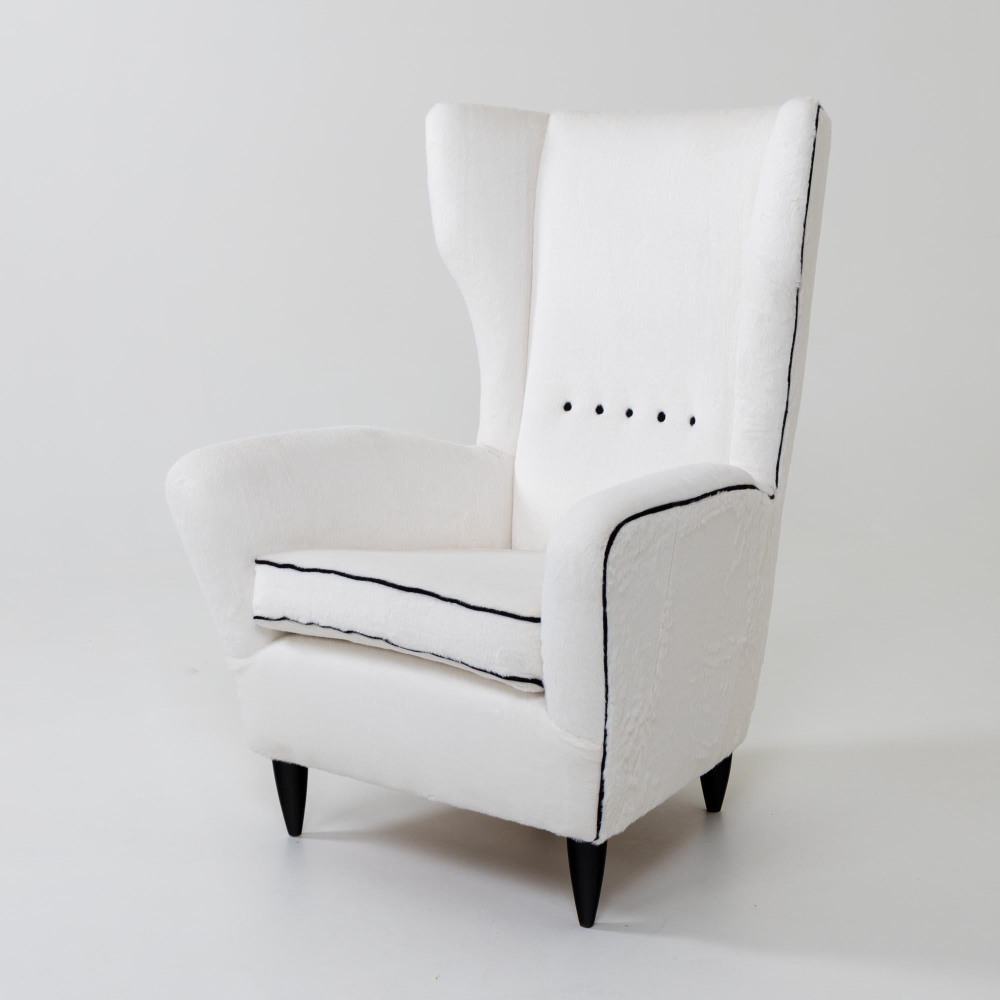 Large wingback armchair on conical black pointed feet. The armchair has been newly upholstered with a high-quality white imitation fur and accentuated with black piping and buttons.
