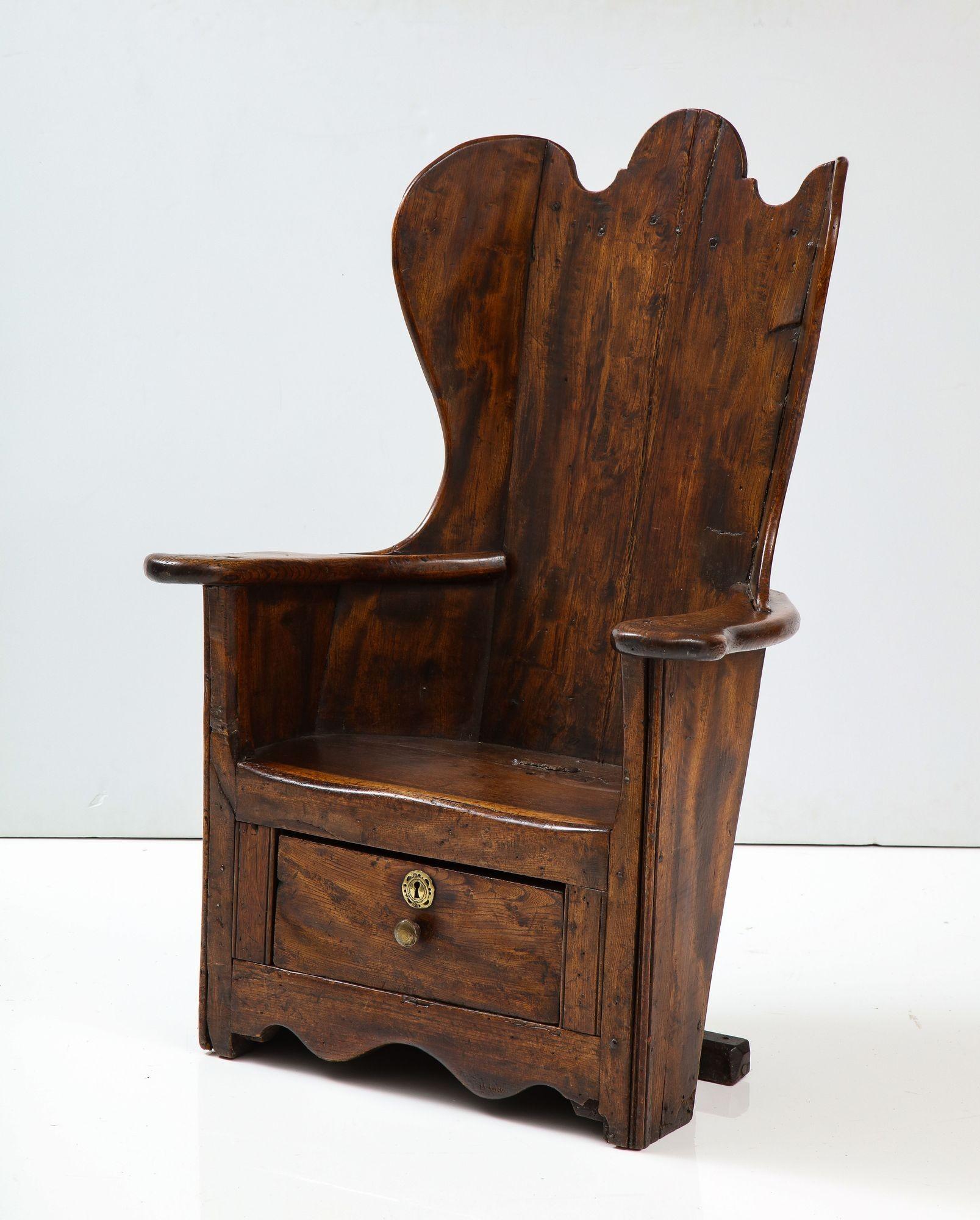 A paneled chair in elm and oak with solid back and sides and single drawer under seat, with well-formed curved flattened arms ending in broadened hand supports suitable for supporting a cup of tea, featuring canted side wings and a deeply scalloped