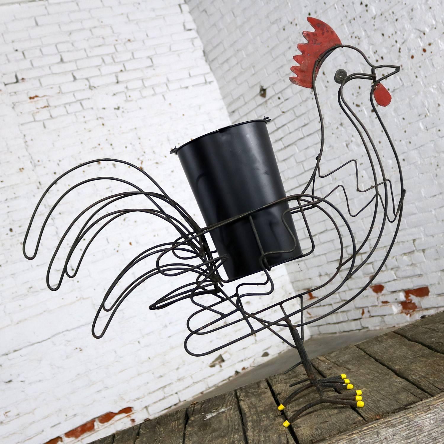 Over-scaled wire rooster planter with a red comb, yellow rubber toes, and a black bucket as planter. It is in purposeful distressed condition, circa 21st century.

This handsome guy is strutting his stuff! He is a quirky rooster planter comprised