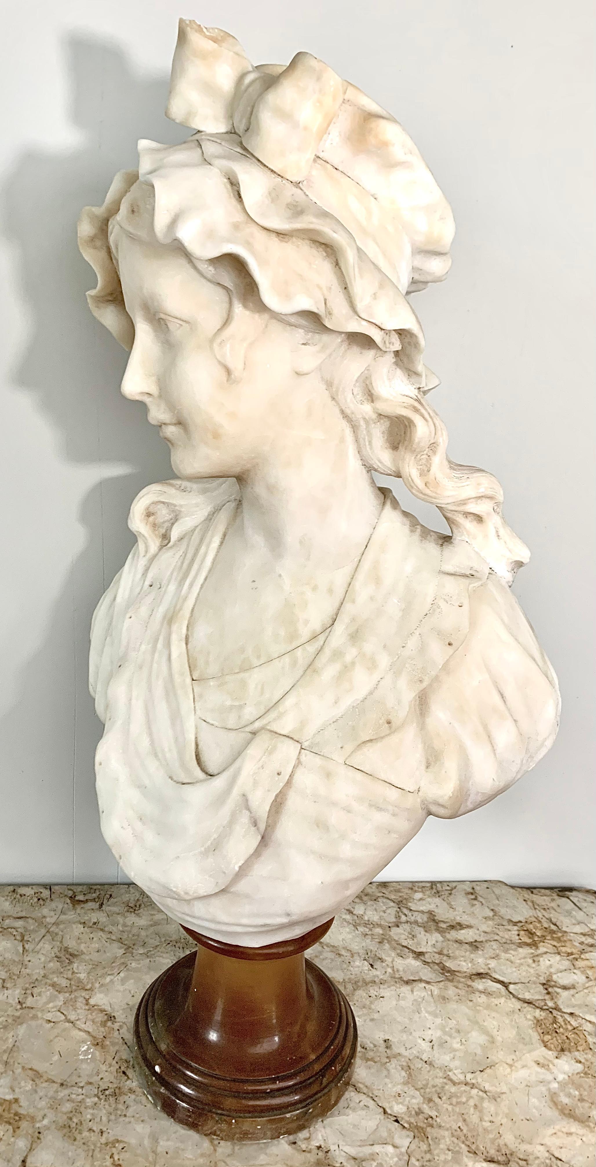 Charming alabaster bust of a young woman. Her head is covered with a small peasant bonnet. On this bonnet, a pretty bow is perfectly sculpted. A few strands of wavy hair are escaping from it. Her face is turned 3/4, giving a certain movement to the