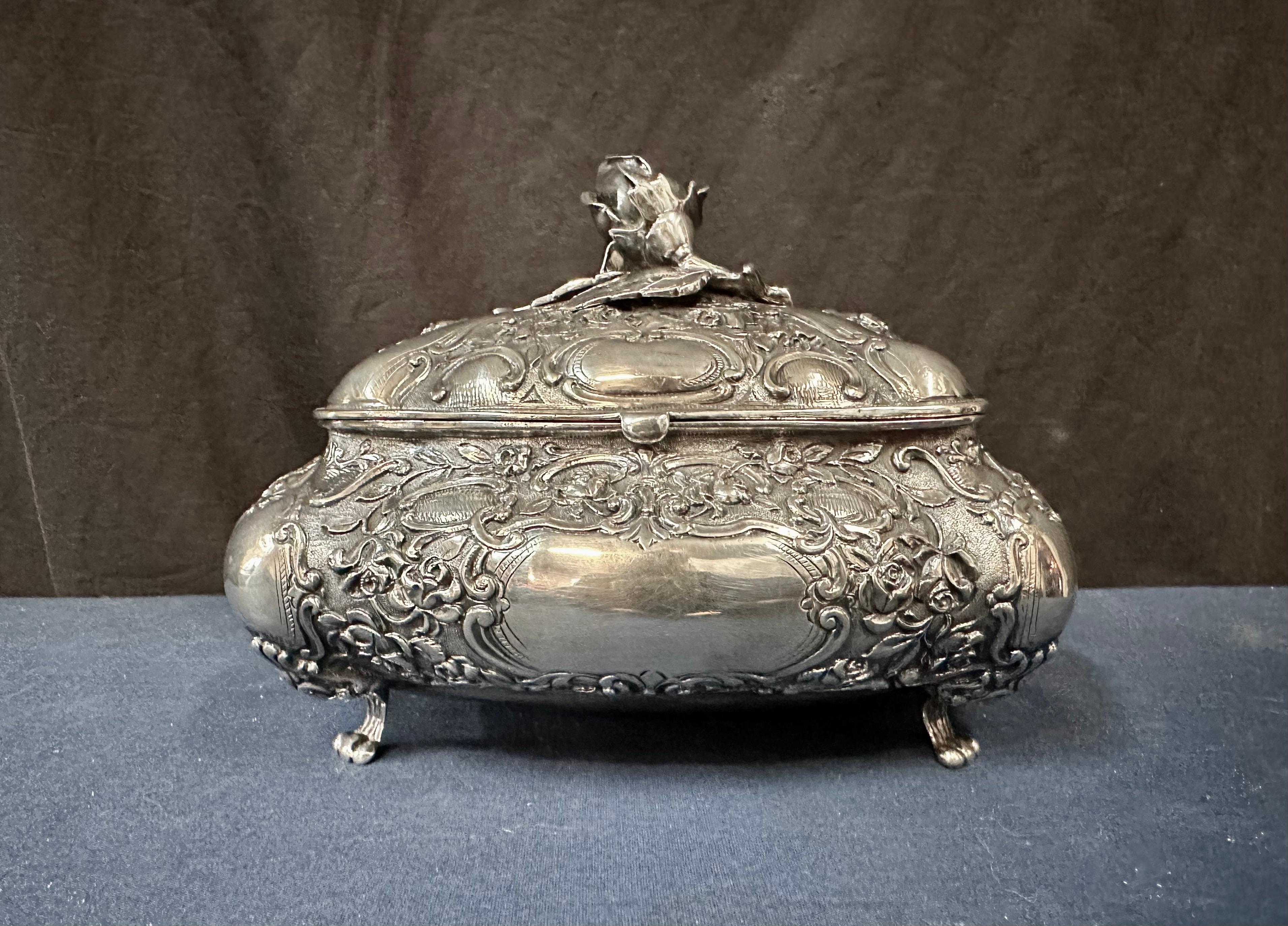 Este vintage alemán de finales del siglo XIX  La caja para el té está diseñada con un magnífico y detallado motivo floral repoussé. Varias enredaderas, hojas y rosas fluyen sobre la caja con pie. Entre la flora, se encuentran escudos pulidos sin