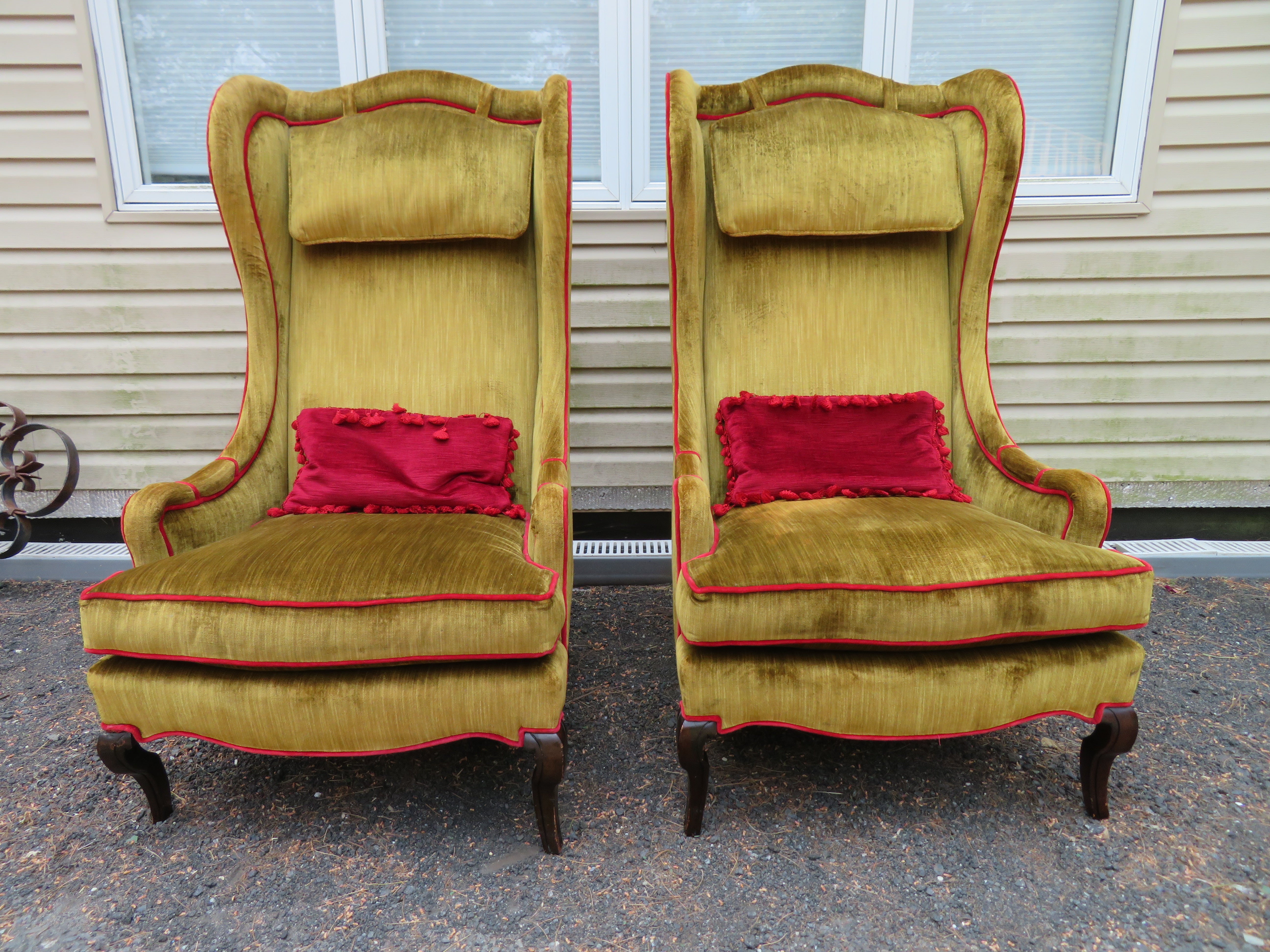 Wonderful pair of Dorothy Draper-style tall wingback chairs. We just love the way the original designer upholstered these with the bold red piping and matching fringed velvet lumbar pillows-very charming indeed. The original olive green velvet shows