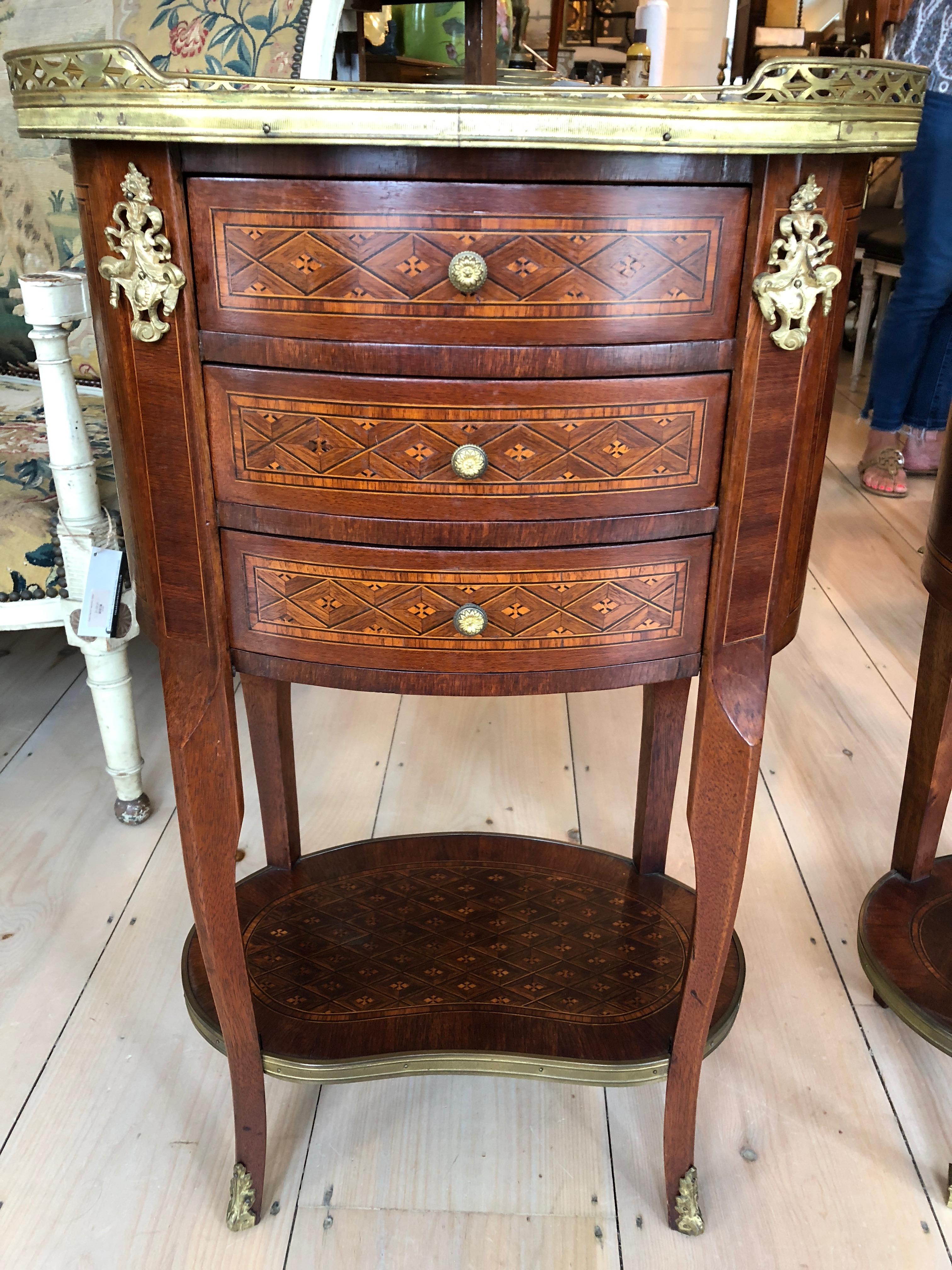 Elegant and unusual to find pair of mahogany oval night stands having creamy marble tops surrounded by brass gallery, three drawers, lovely inlay work, and ormolu details. The best part is that they are not exactly alike, with small variations that