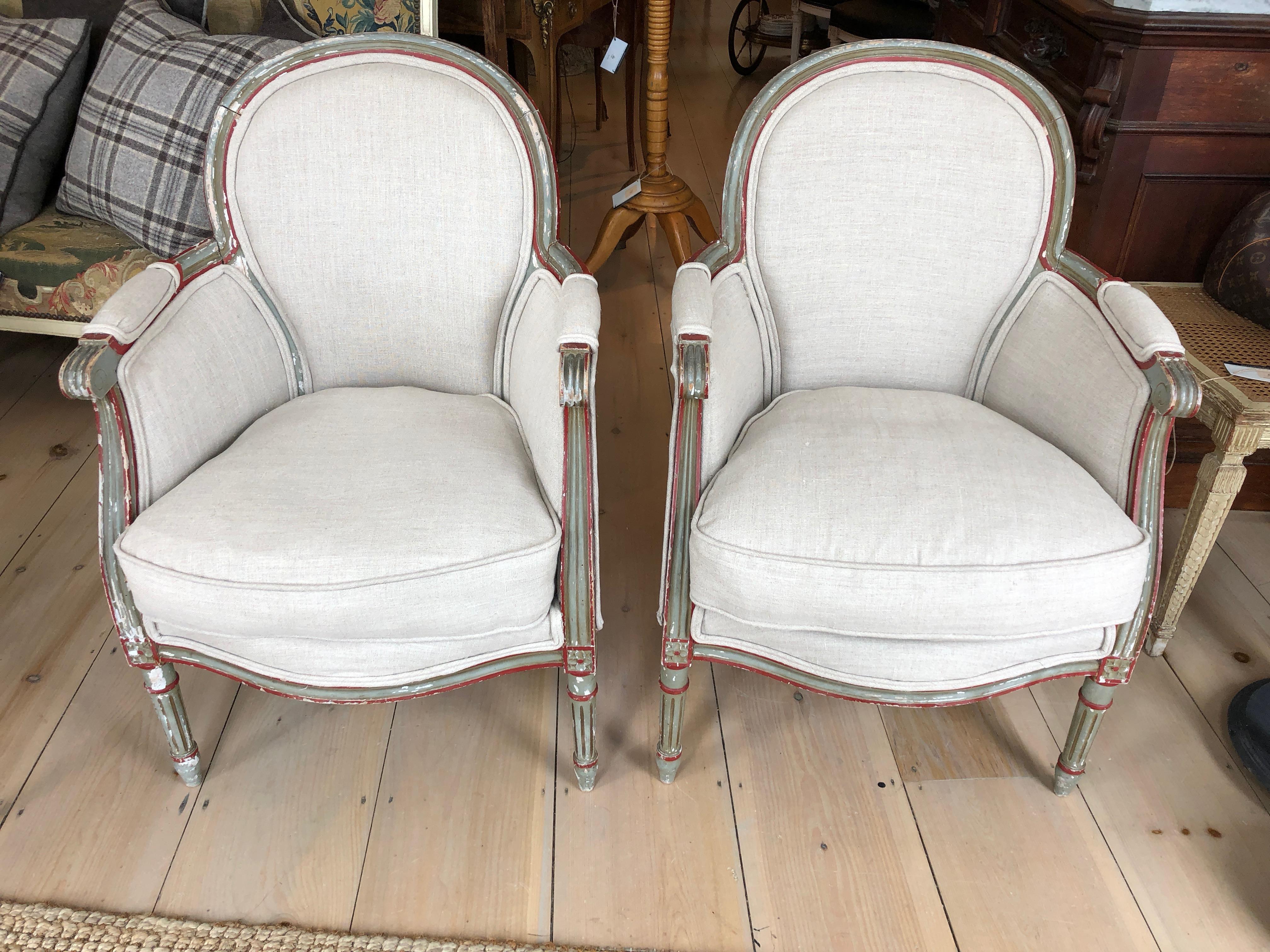 A charming diminutive apartment sized pair of vintage bergeres having painted wood frames in wonderful contrasting hookers green and red oxide, newly upholstered in neutral beige linen. Window pane backs and chippy paint here and there add oodles of