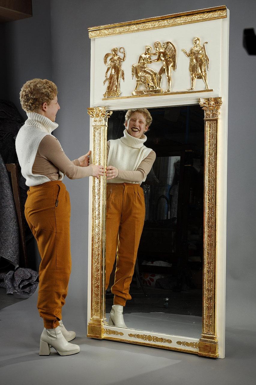 Miroir trumeau d'époque Empire, en bois et stuc doré à la feuille d'or. Ce miroir est encadré par deux pilastres de style antique en bois et stuc, dorés à la feuille d'or. Un fronton rectangulaire rehausse l'ensemble et accueille une scène