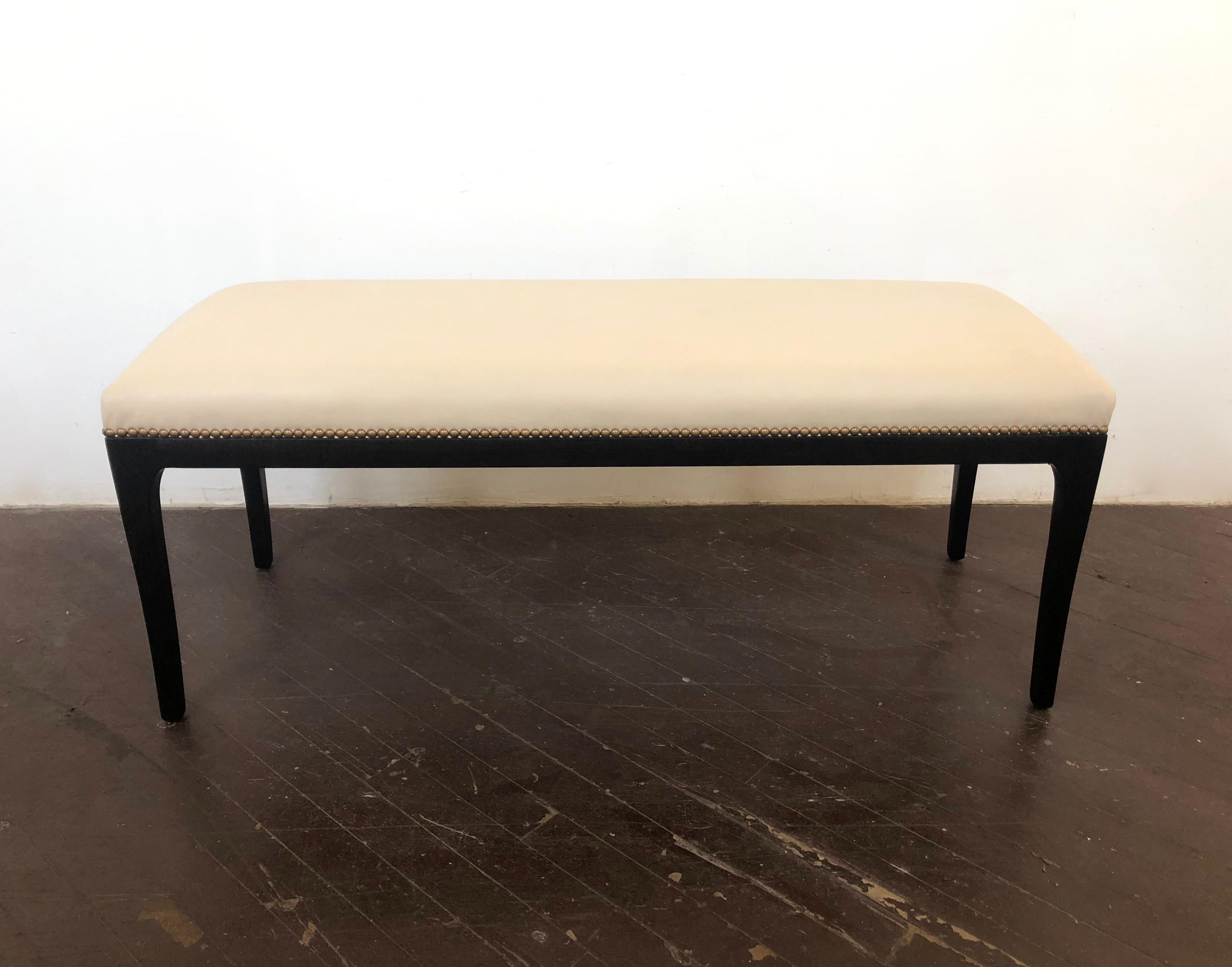 1950s restored wooden framed bench with rounded corners and four slightly tapered legs that support a spring backed seat cushion that has been reupholstered in cream colored leather and brass nailheads along the border.