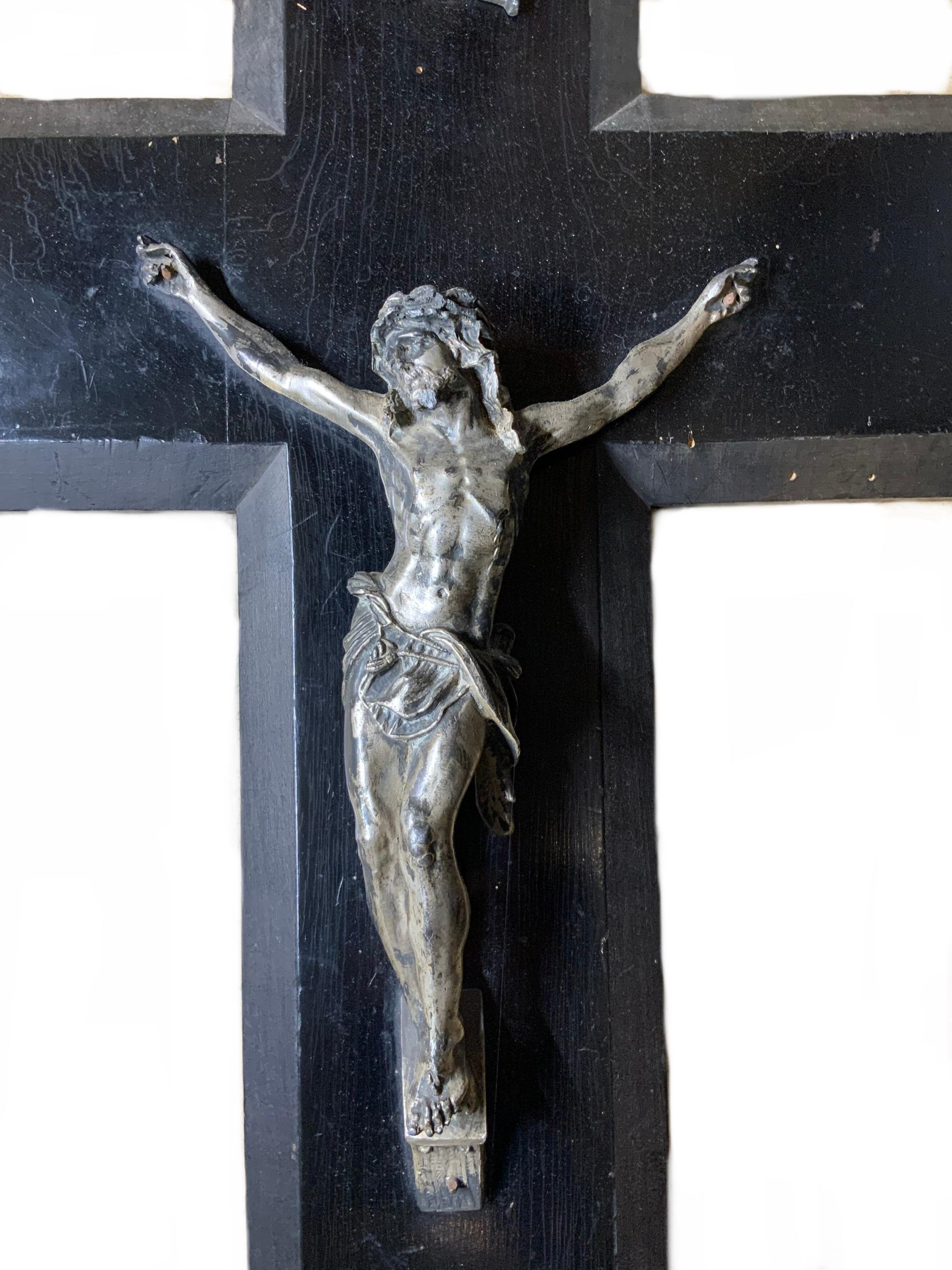This is a wall crucifix benetier made of wood and metal. The wood cross is hand painted black. The Jesus Christ and the blessing bowl are made of silver tone metal that is adorned with embossed foliage. Above the Jesus Christ there is a scroll with